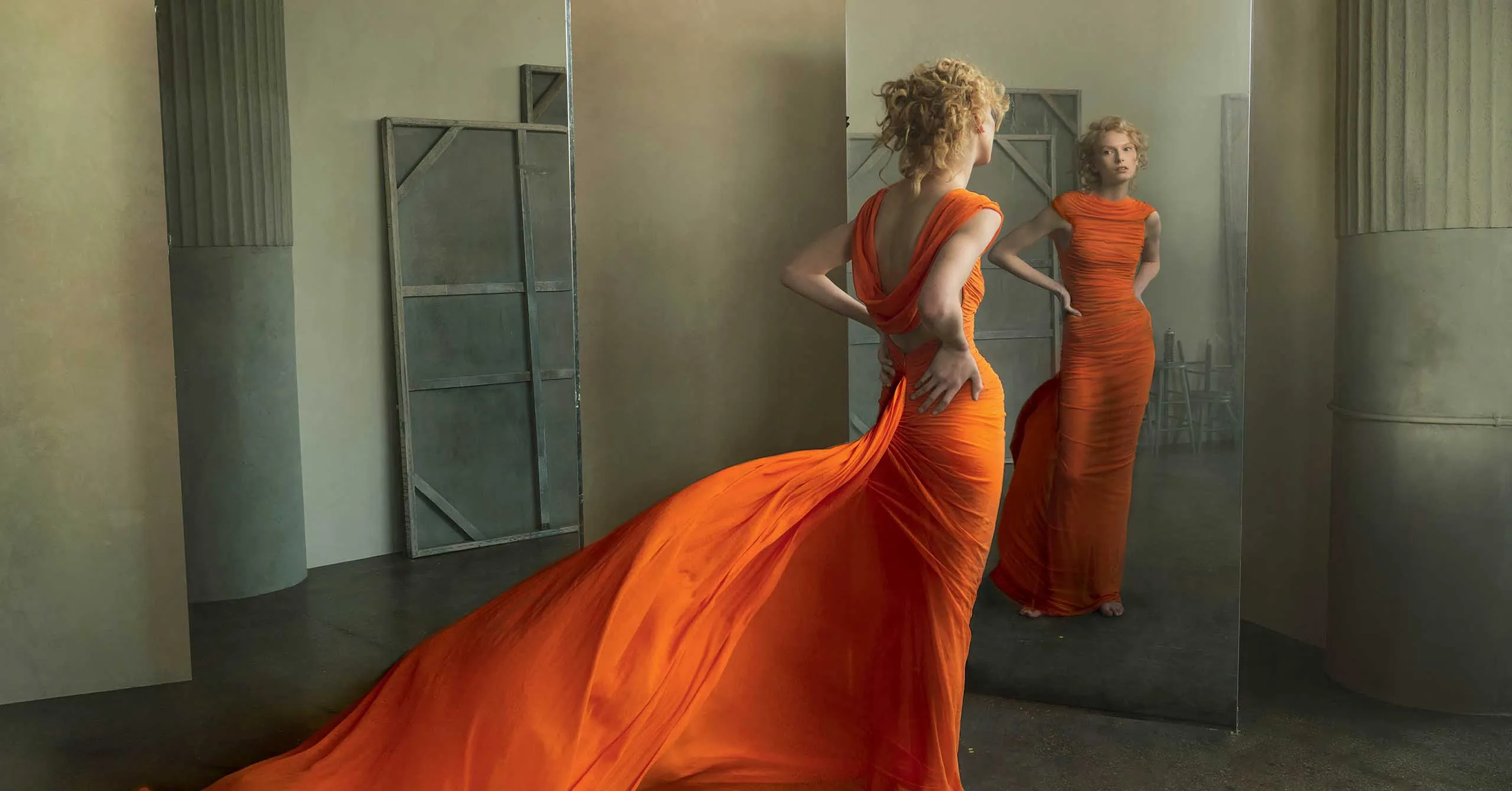 Model wearing a bright orange gown with a long train, posing before a mirror in a muted industrial studio.