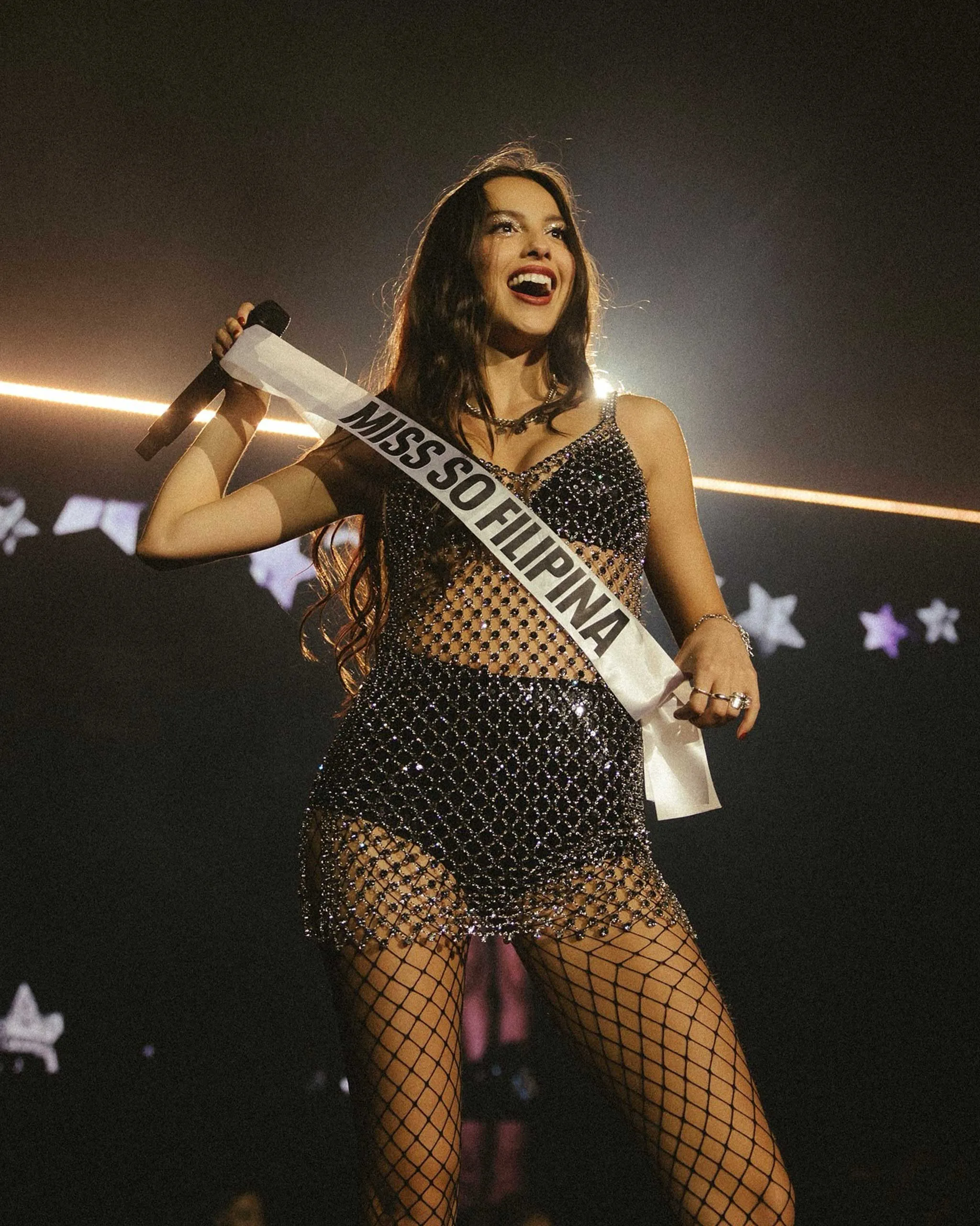 Miss So Filipina contestant on stage, smiling with a microphone, wearing a beaded fishnet dress and sash with 'MISS SO FILIPINA' text.