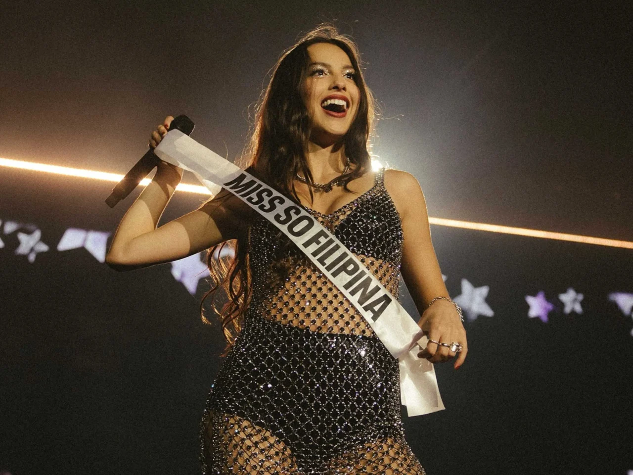 Miss Sofia pageant contestant on stage, smiling in a sequined gown with a sash reading MISS SOFIA, microphone in hand.