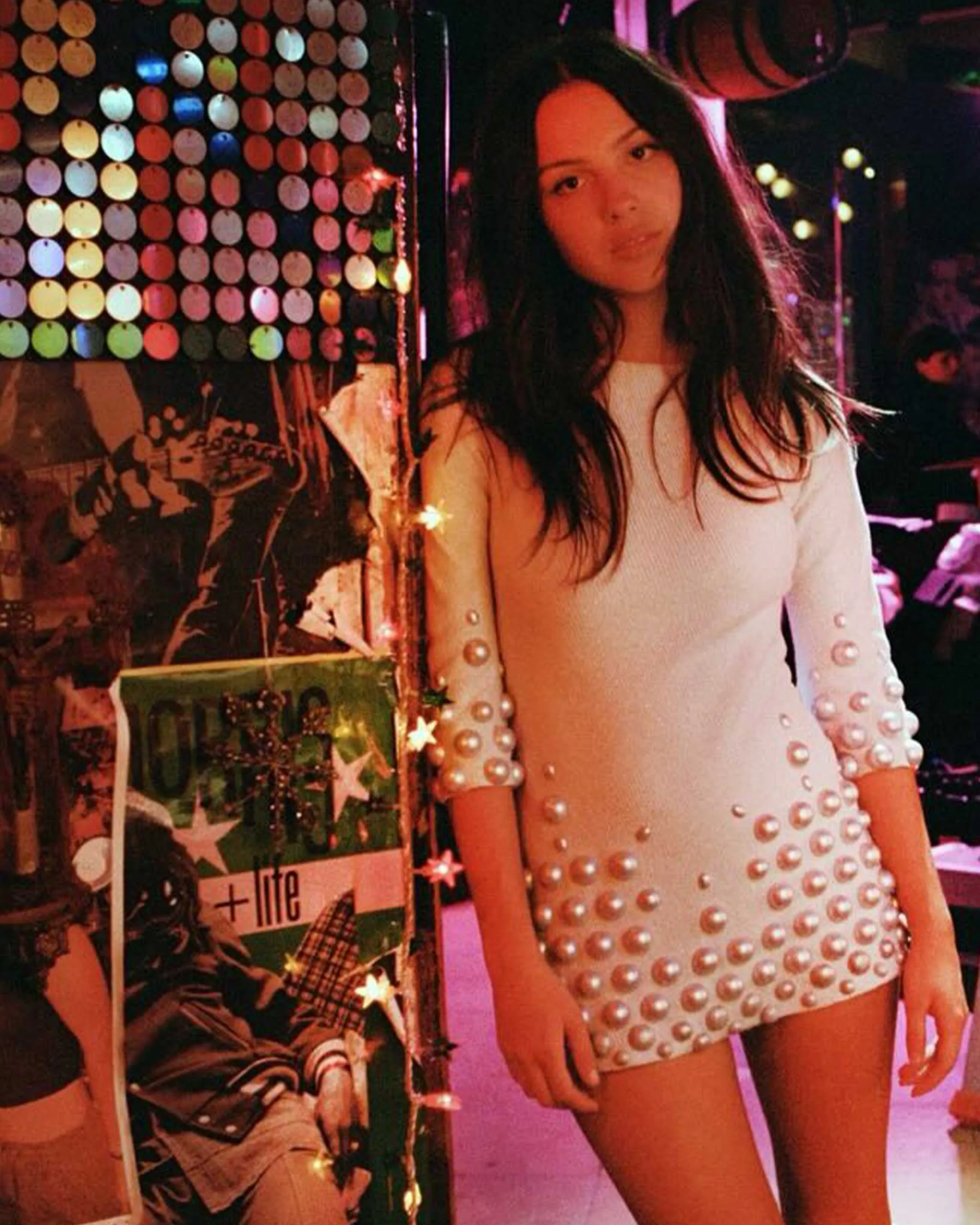 Young woman in a white, pearl-embellished dress posing in a colorful nightclub with neon lights.