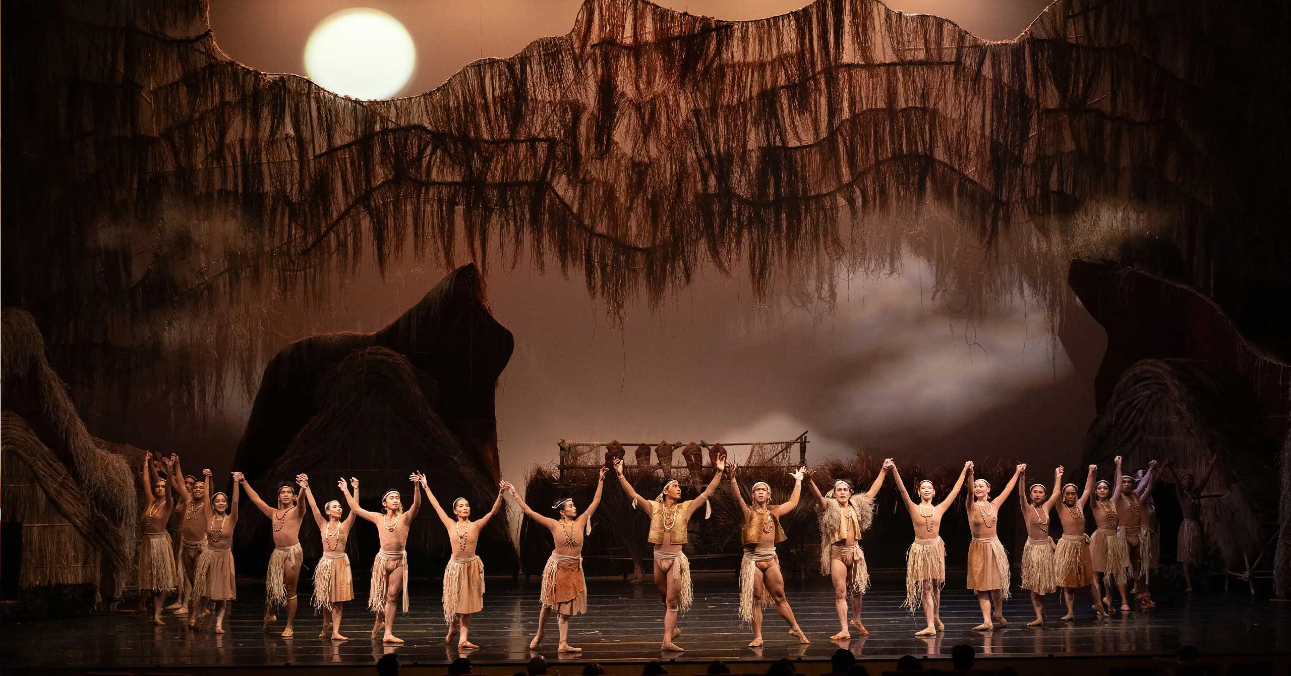 Dancers in tan grass-skirt costumes hold hands overhead in a line on a stage with a thatched backdrop and a large setting sun behind them.