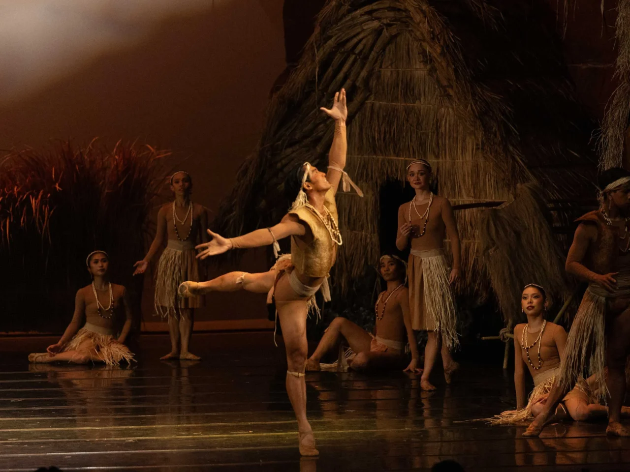 Dancers in tan grass-skirt costumes hold hands overhead in a line on a stage with a thatched backdrop and a large setting sun behind them.