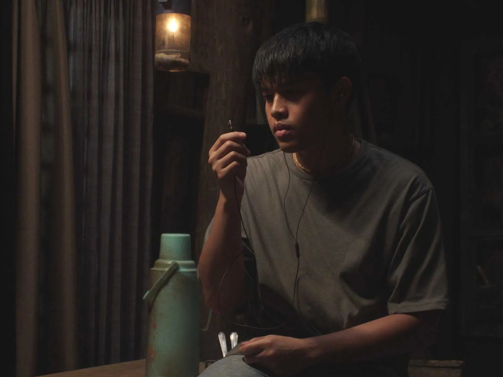 Dimly lit room with a young man in a gray shirt wearing earphones, holding a small object near his face beside a green thermos on a table.