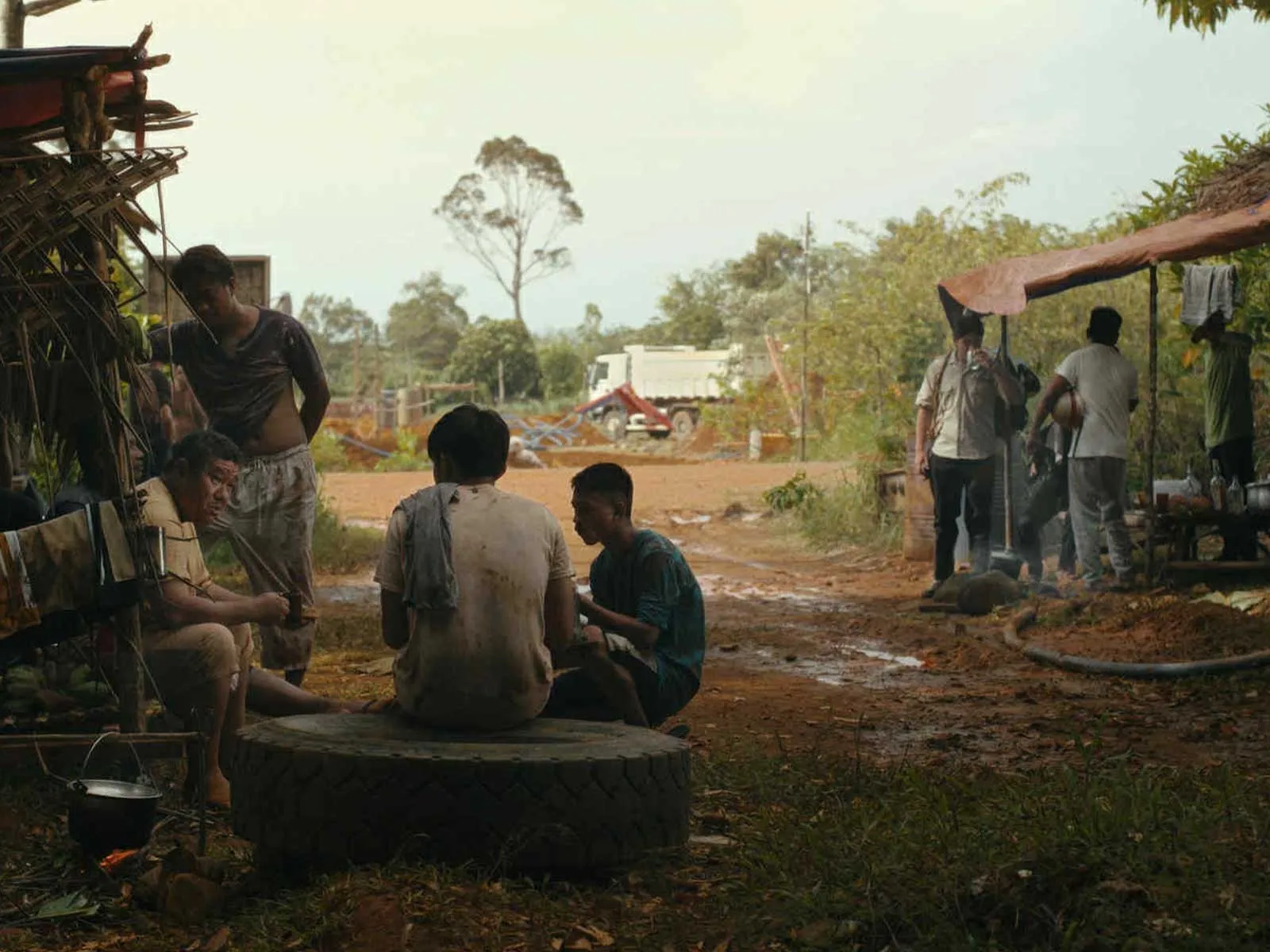 Group of men sits on a large tire, chatting around a makeshift outdoor work site with a cooking pot nearby in a dusty rural setting.