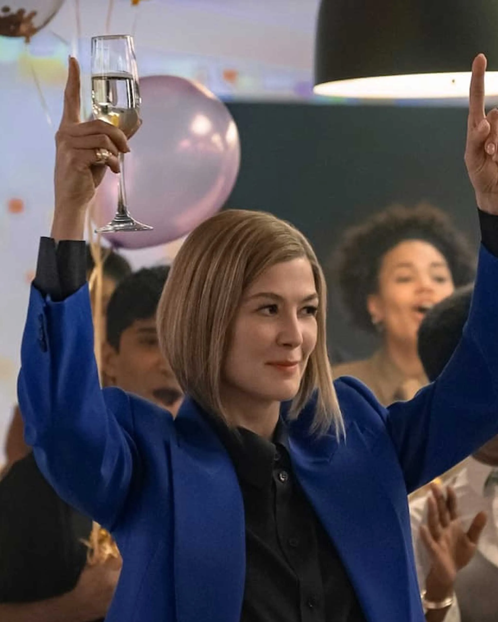 Person in a blue blazer raises a glass of champagne at a celebration, with balloons in the background.