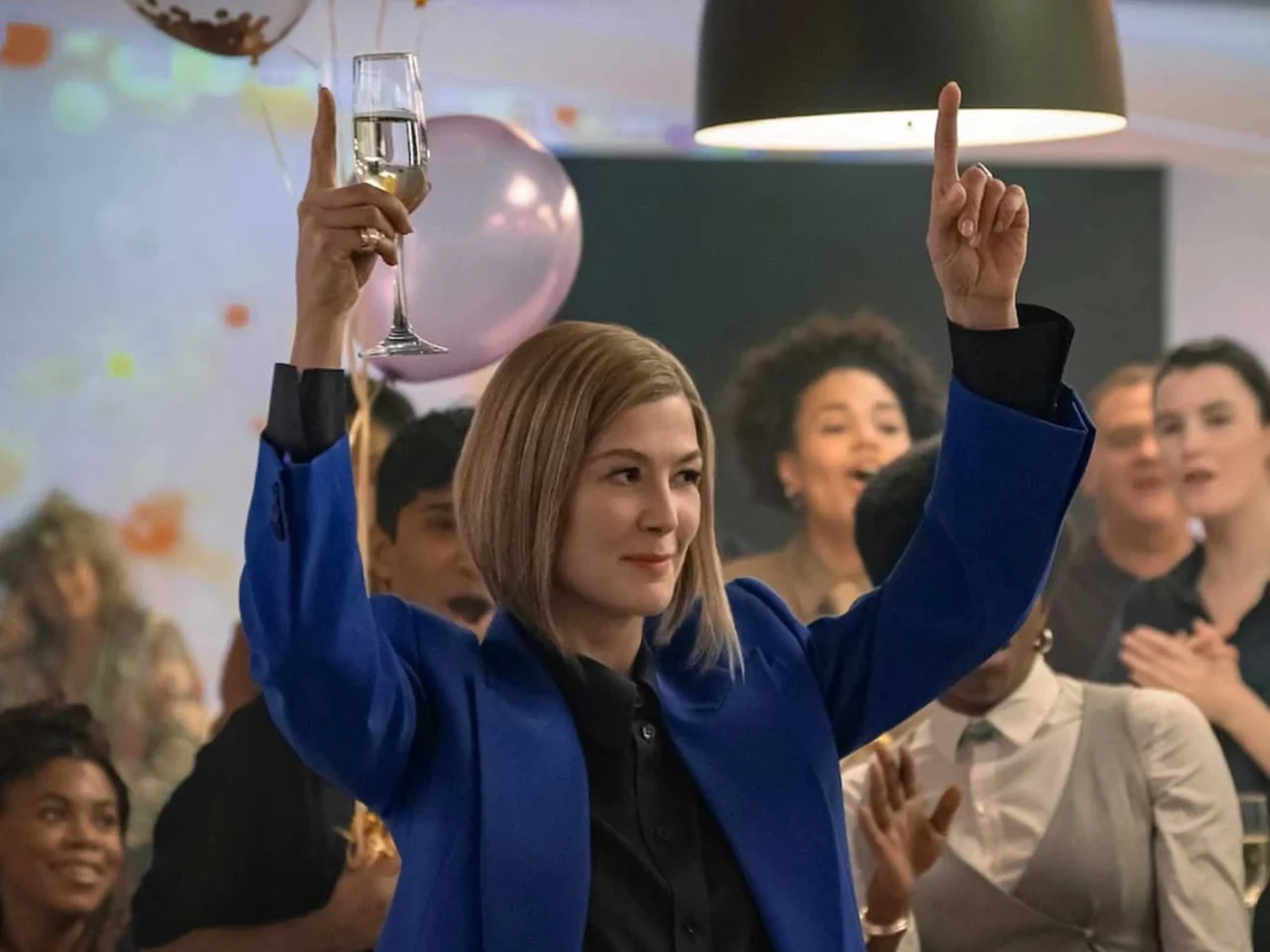 Woman in a blue blazer raises a champagne flute at a celebration with a cheering crowd behind her.