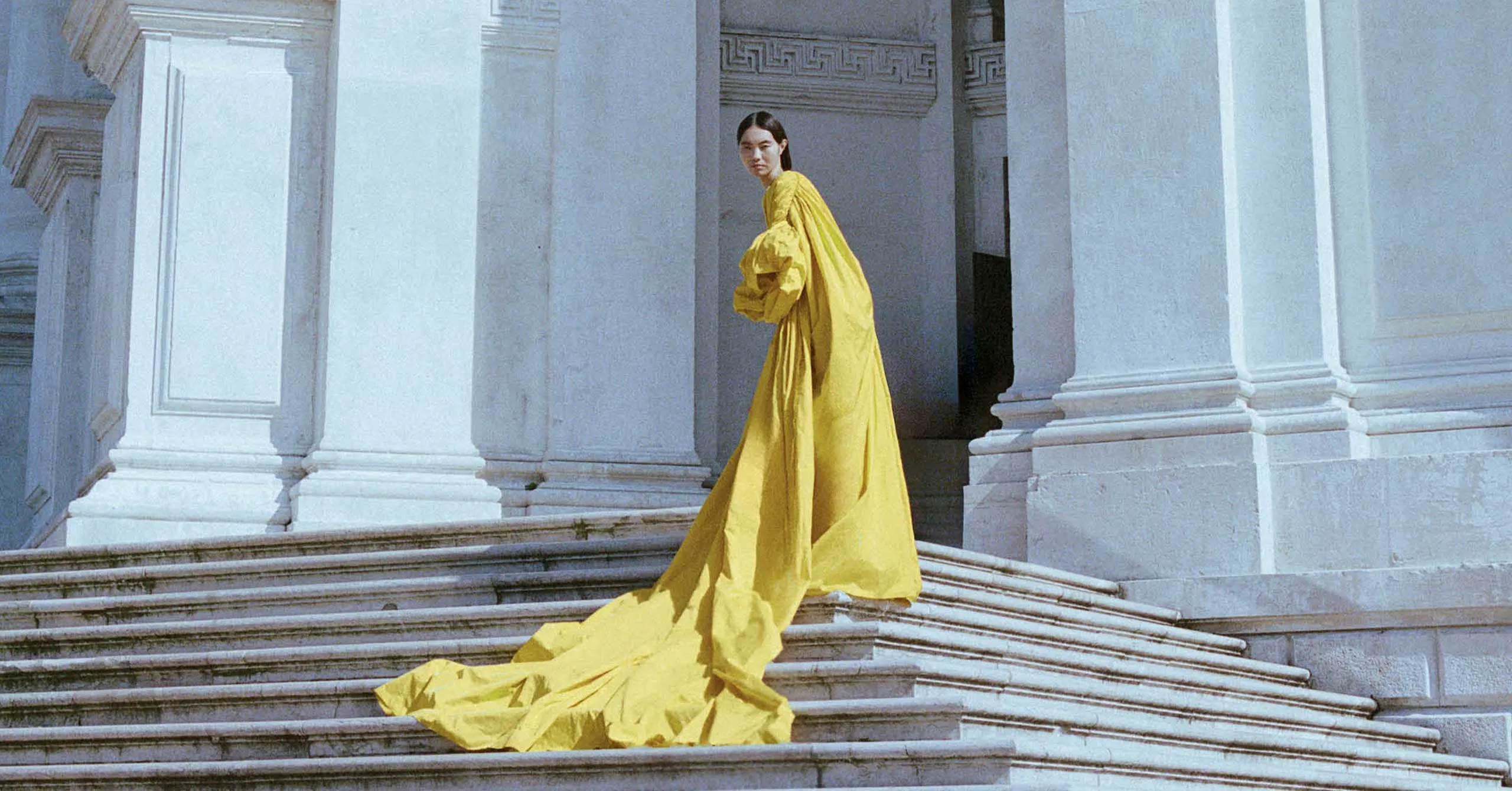 Woman in a flowing bright yellow gown stands on white marble steps of a neoclassical building, looking back over her shoulder