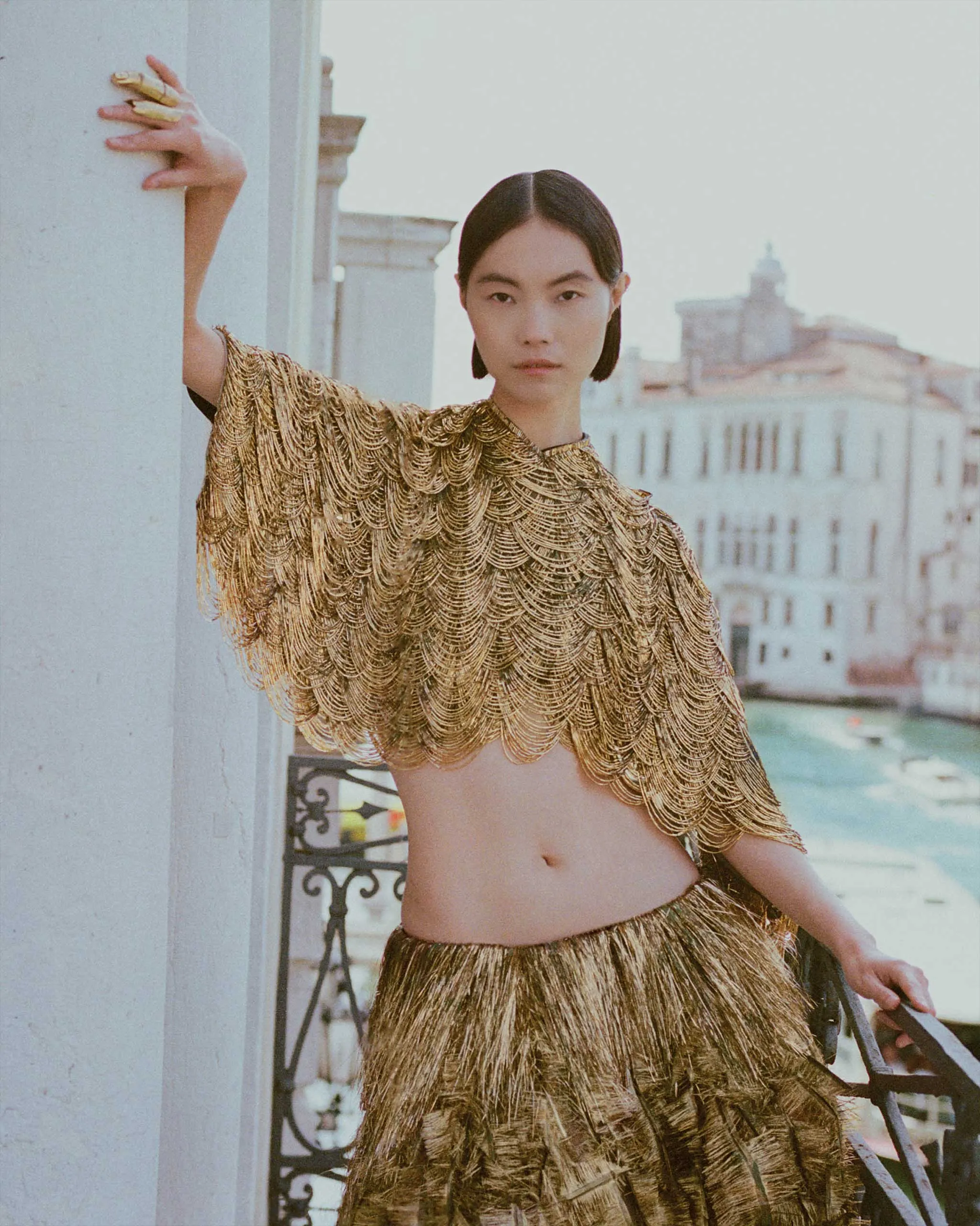 Woman wearing a shimmering gold fringe top leans against a light-colored wall on a balcony, city buildings visible in the background.