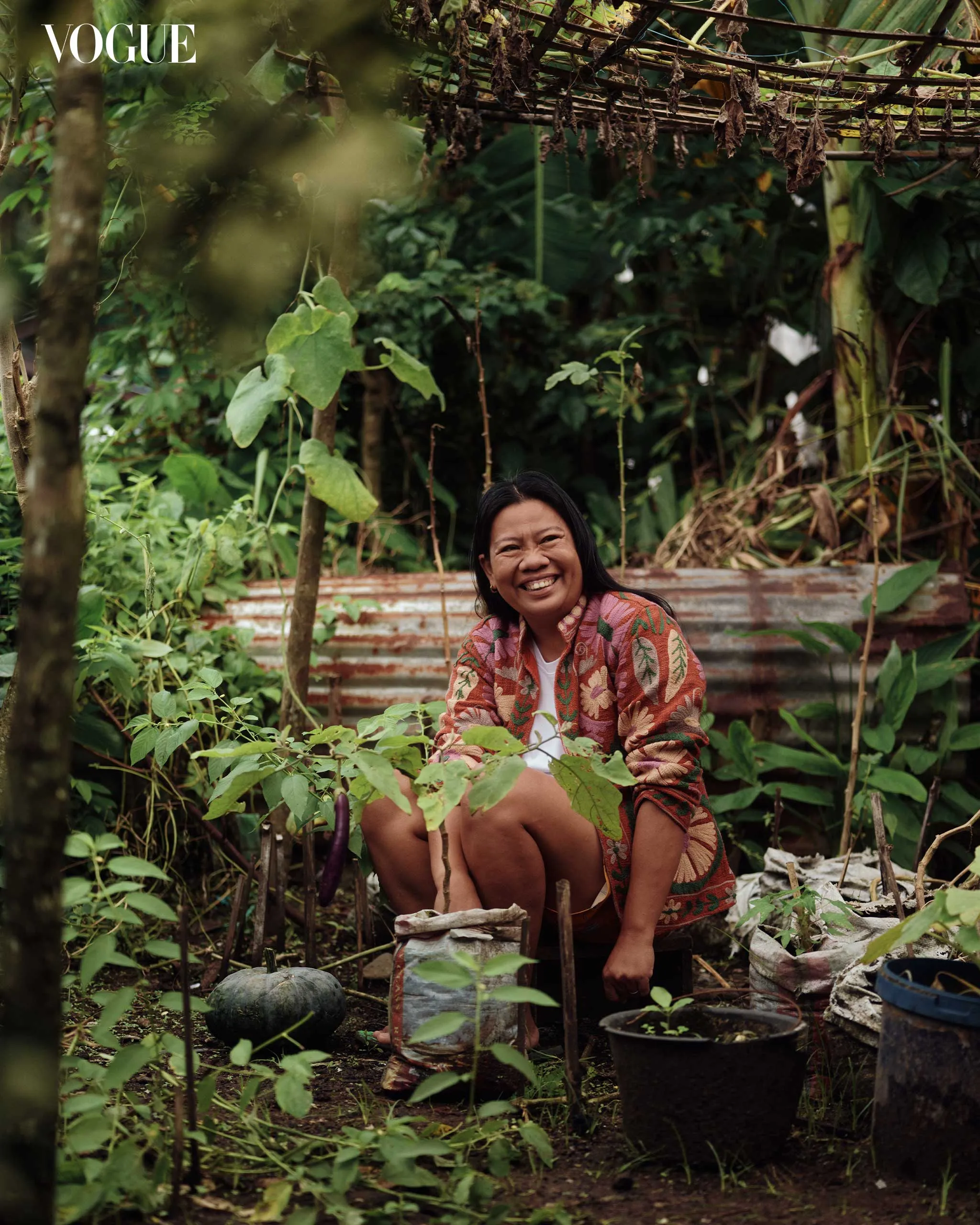 Smiling woman sits among lush garden plants with a patterned shawl, Vogue logo visible in the top-left corner.