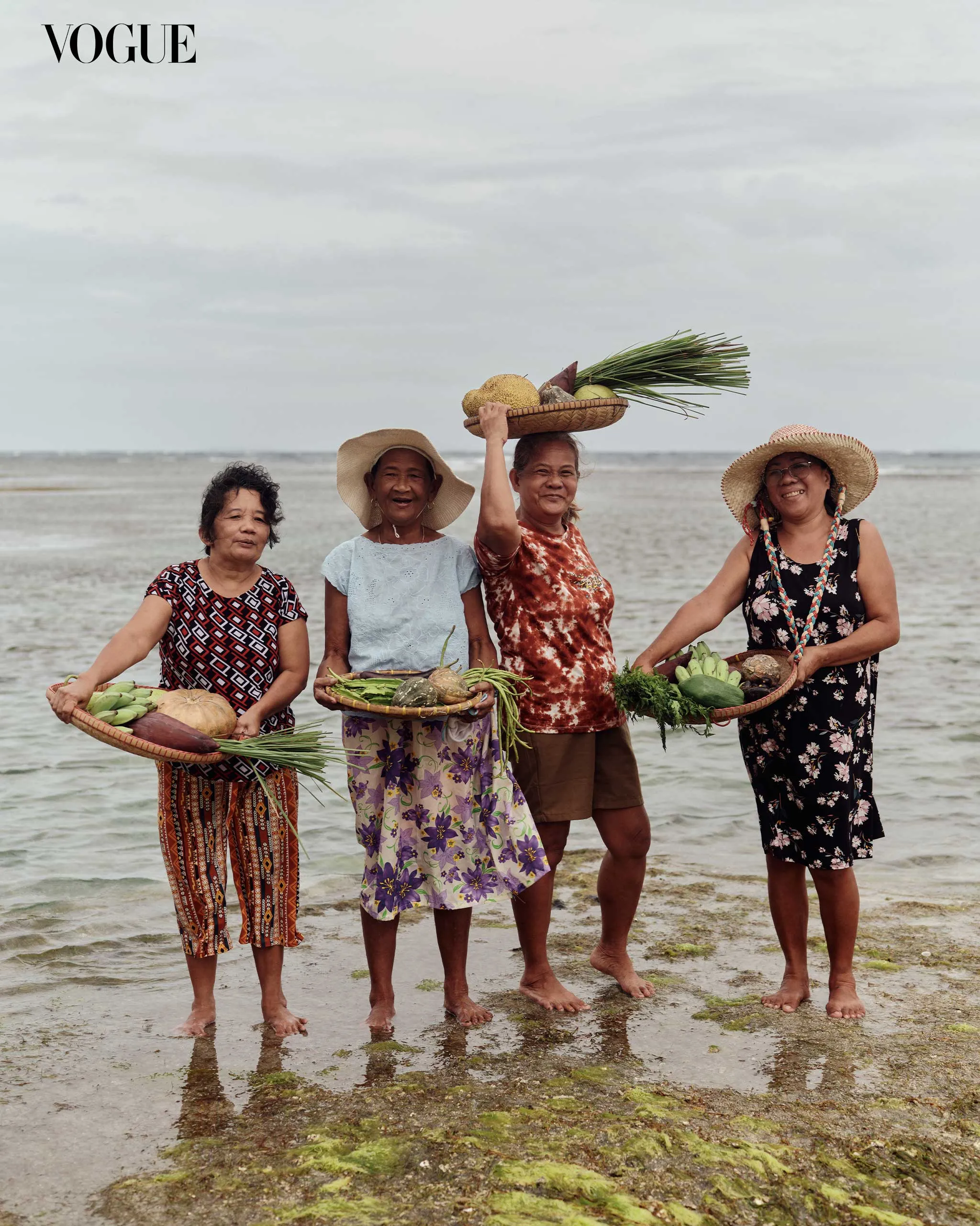 Four women barefoot on a beach, holding baskets of vegetables and wearing sun hats, Vogue logo top-left.