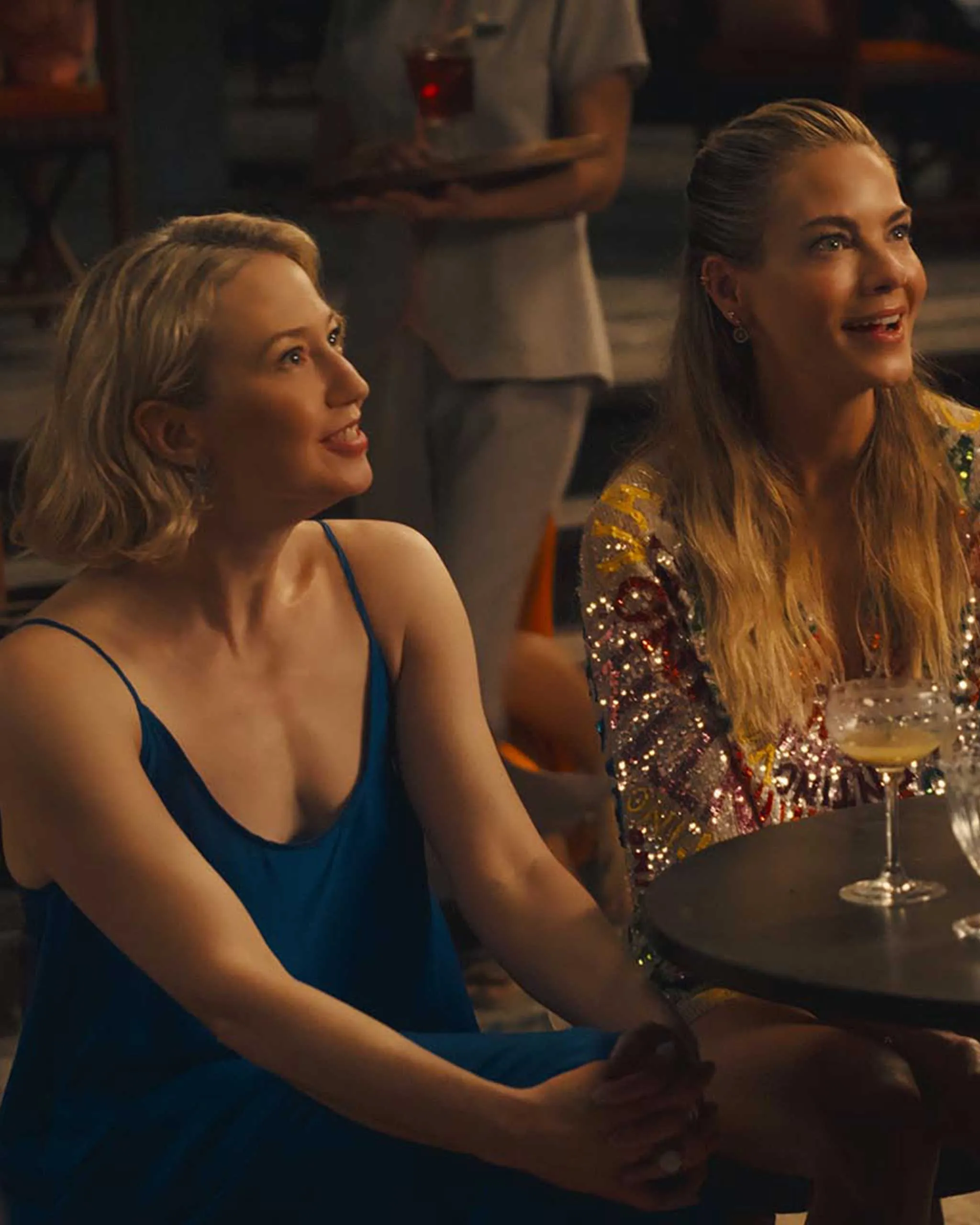 Two women in evening wear chat and smile at a bar with cocktails in front of them, in a dimly lit setting.