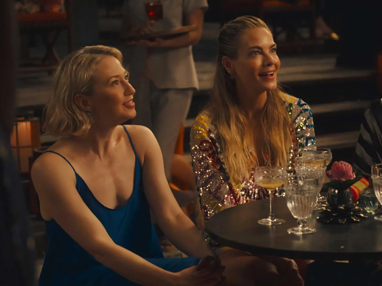 Three women sit at a bar smiling and chatting, with cocktails on the table in front of them.