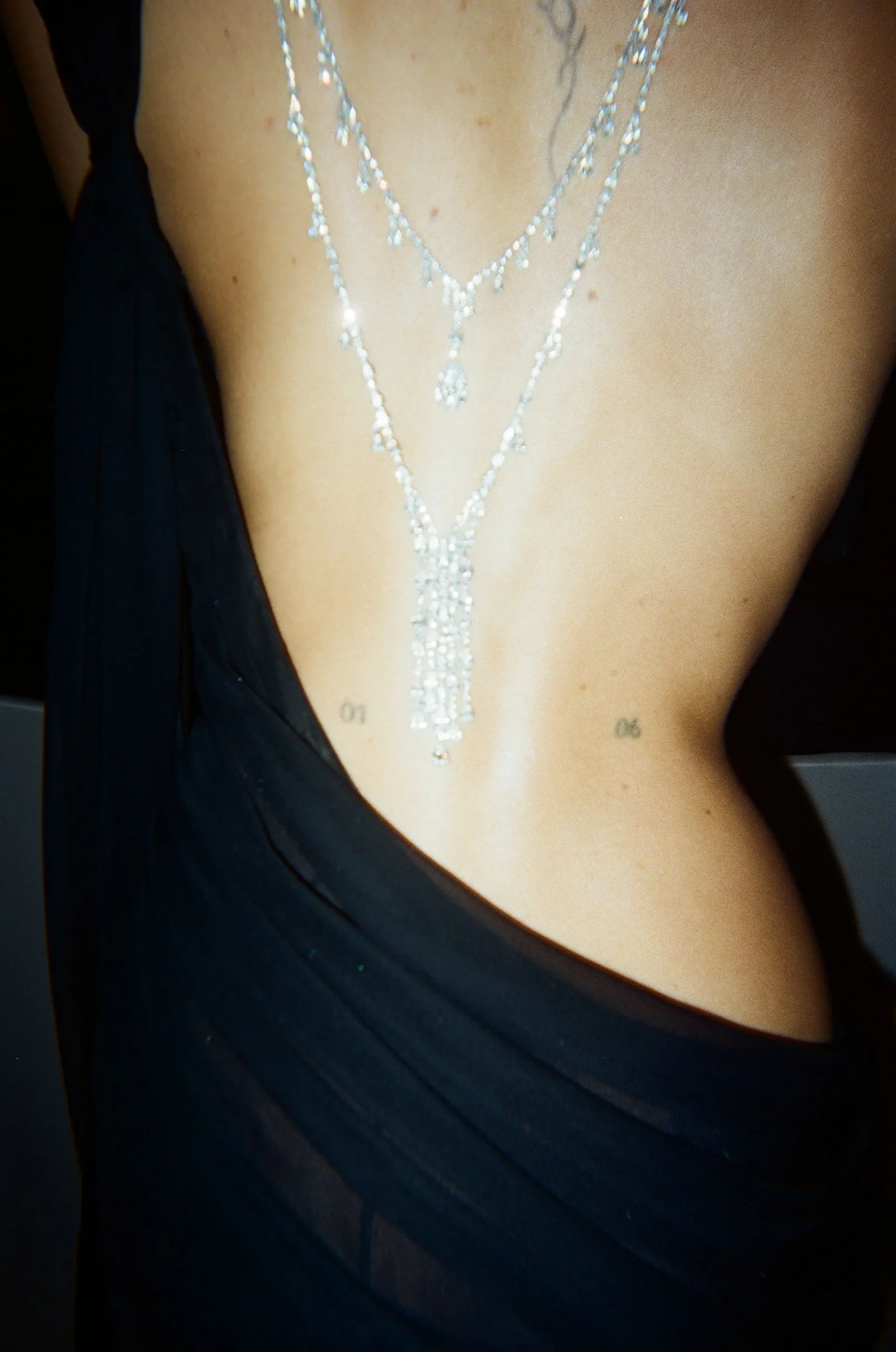 Back of a person in a black dress wearing a layered diamond necklace; small tattoos with '01' and '06' are visible on the lower back.