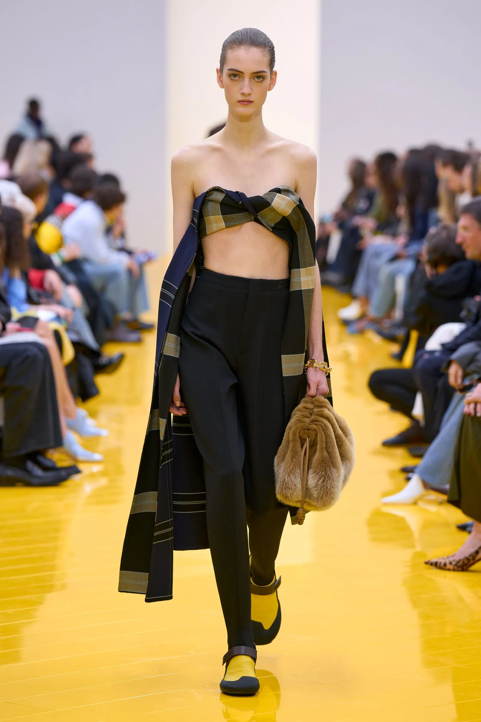 Model walking the runway in a strapless black outfit with a plaid scarf wrapped around the chest, black trousers, and yellow-accent shoes on a yellow catwalk.