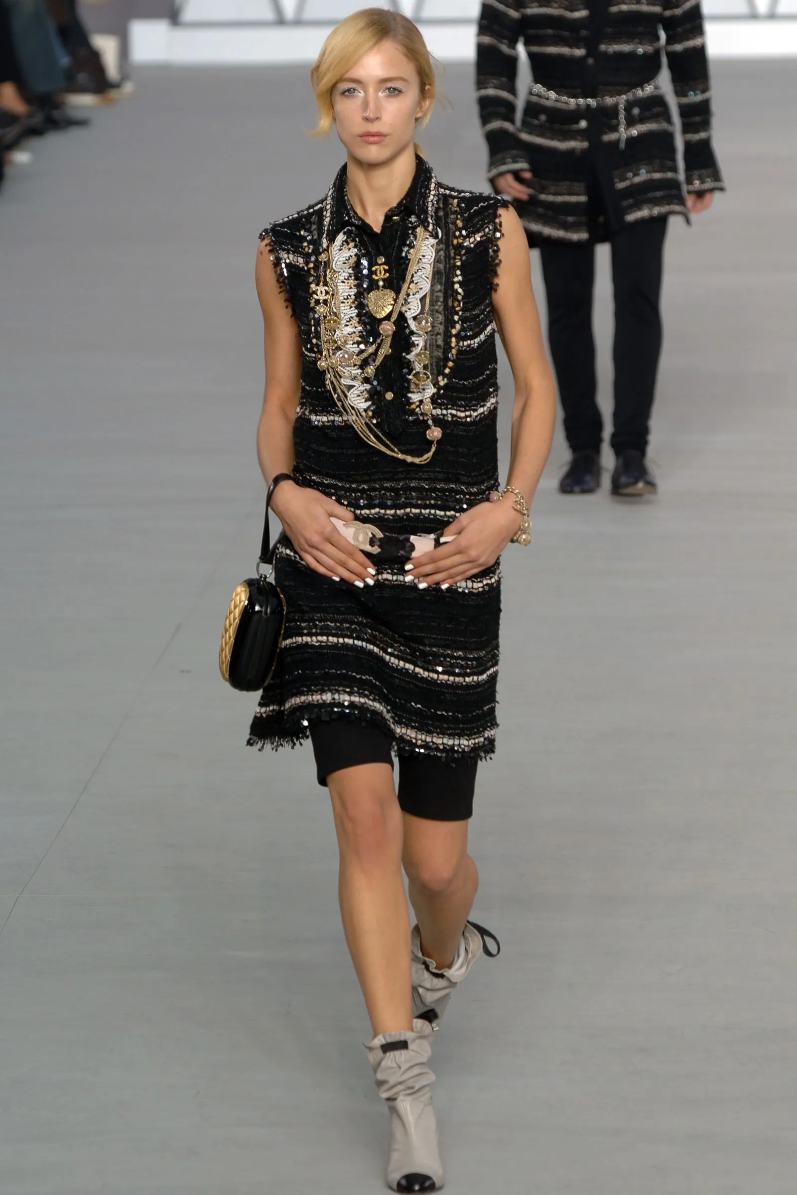 Runway model wearing a black sequined sleeveless dress with gold jewelry and a small handbag.