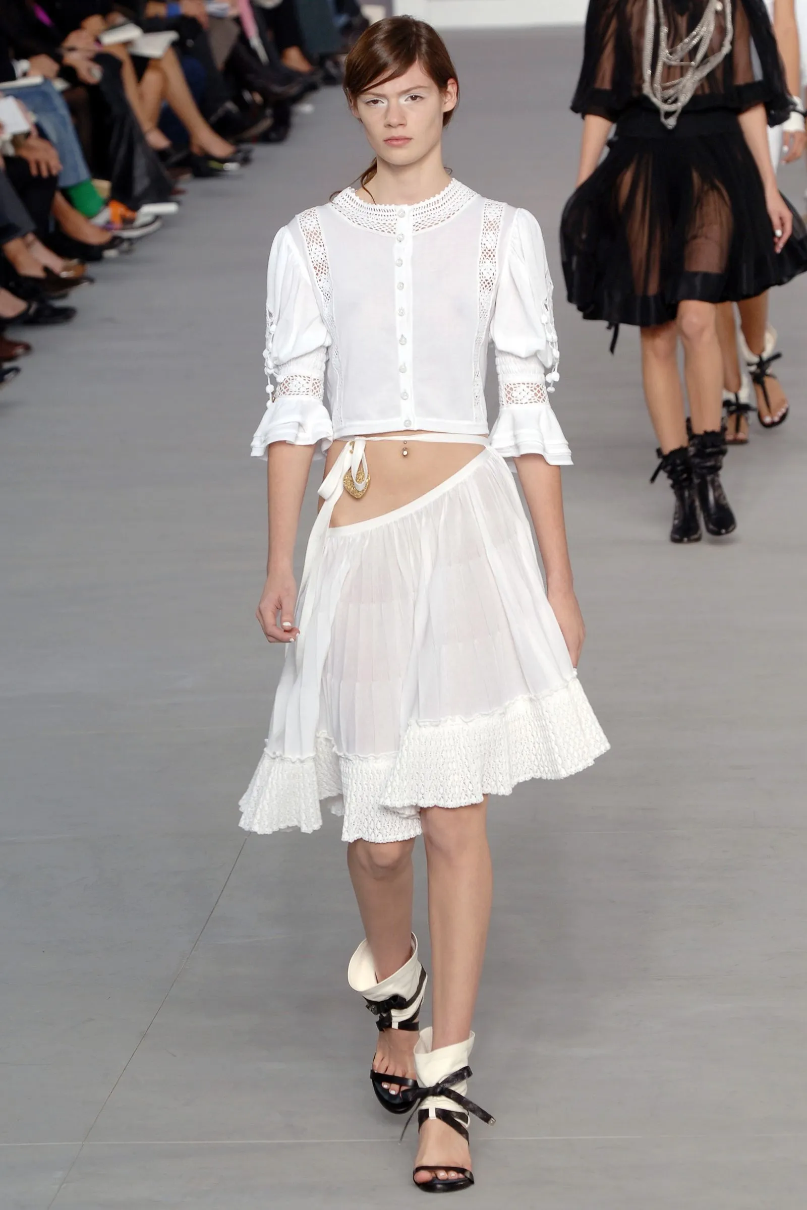 Model walking a runway in a white, lace-trimmed blouse and matching skirt with layered hem and open-toed sandals.