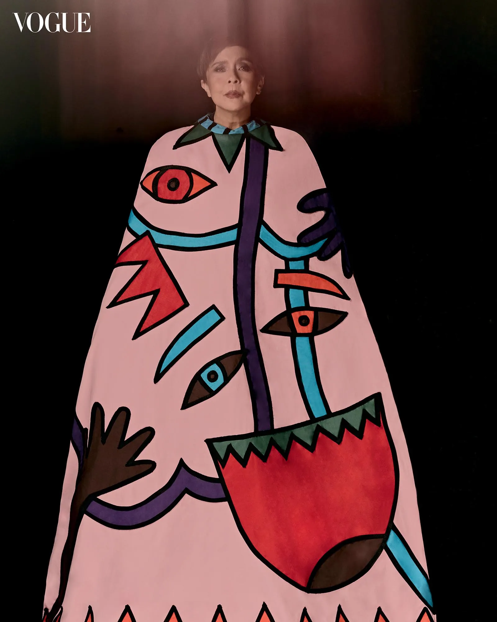 Person wearing a pink, abstract-patterned coat with bold geometric shapes and eye motifs, standing against a dark background with a Vogue watermark in the corner.
