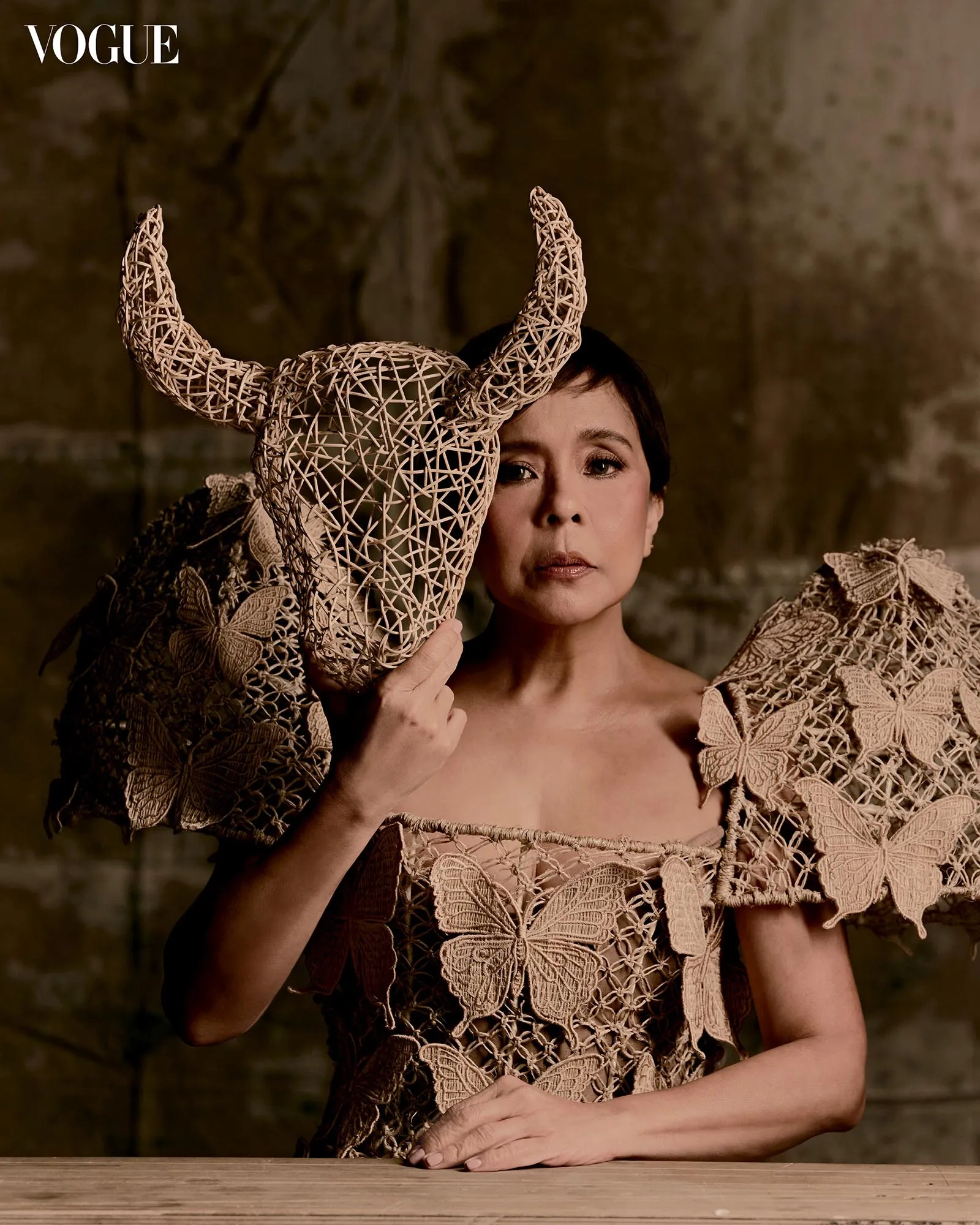 Woman wearing a lace dress holds a large woven wicker headpiece beside her face, Vogue logo in the corner.