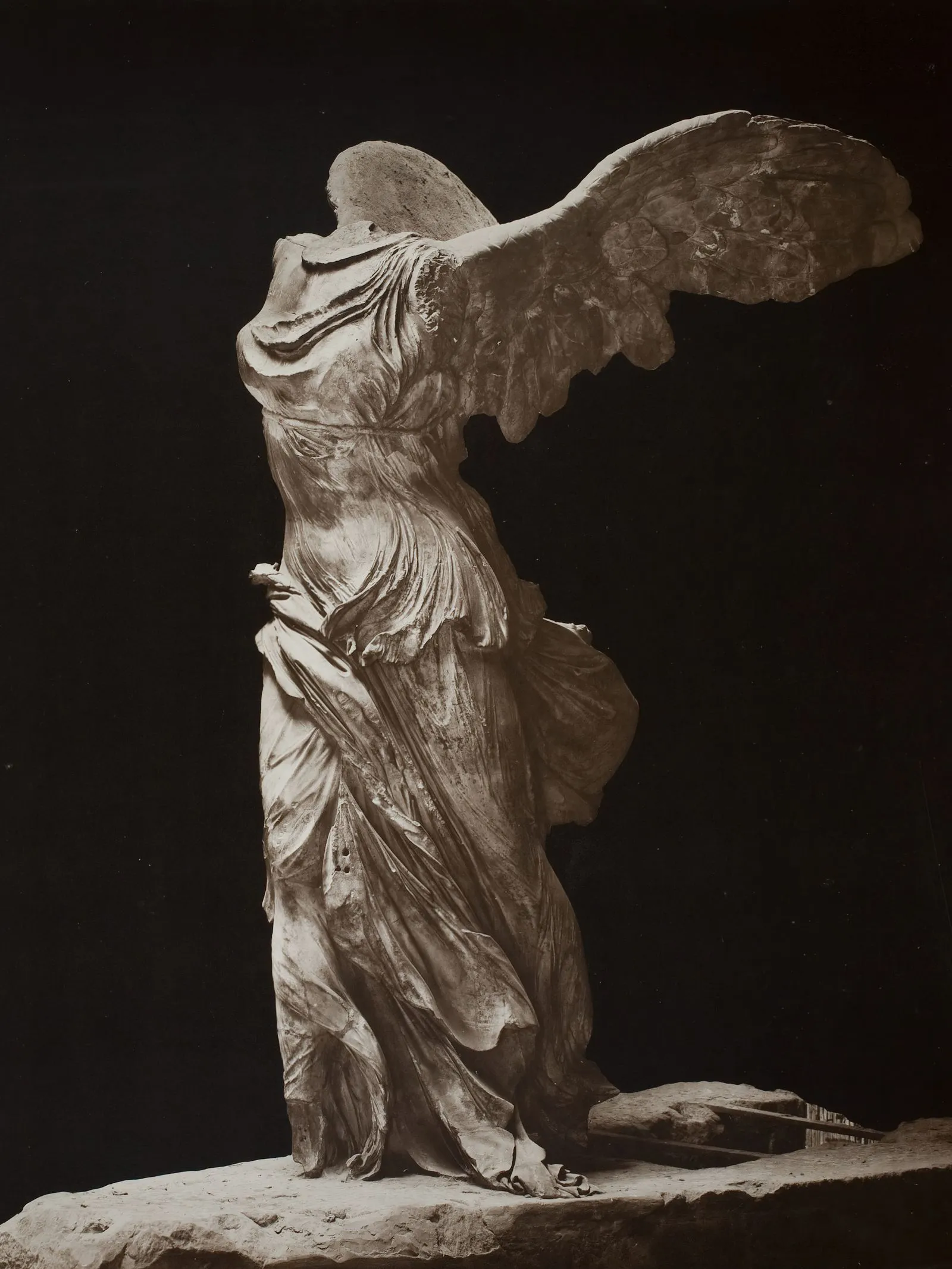Winged ancient statue of a robed female figure standing on a stone pedestal, head and arms missing, against a dark background.