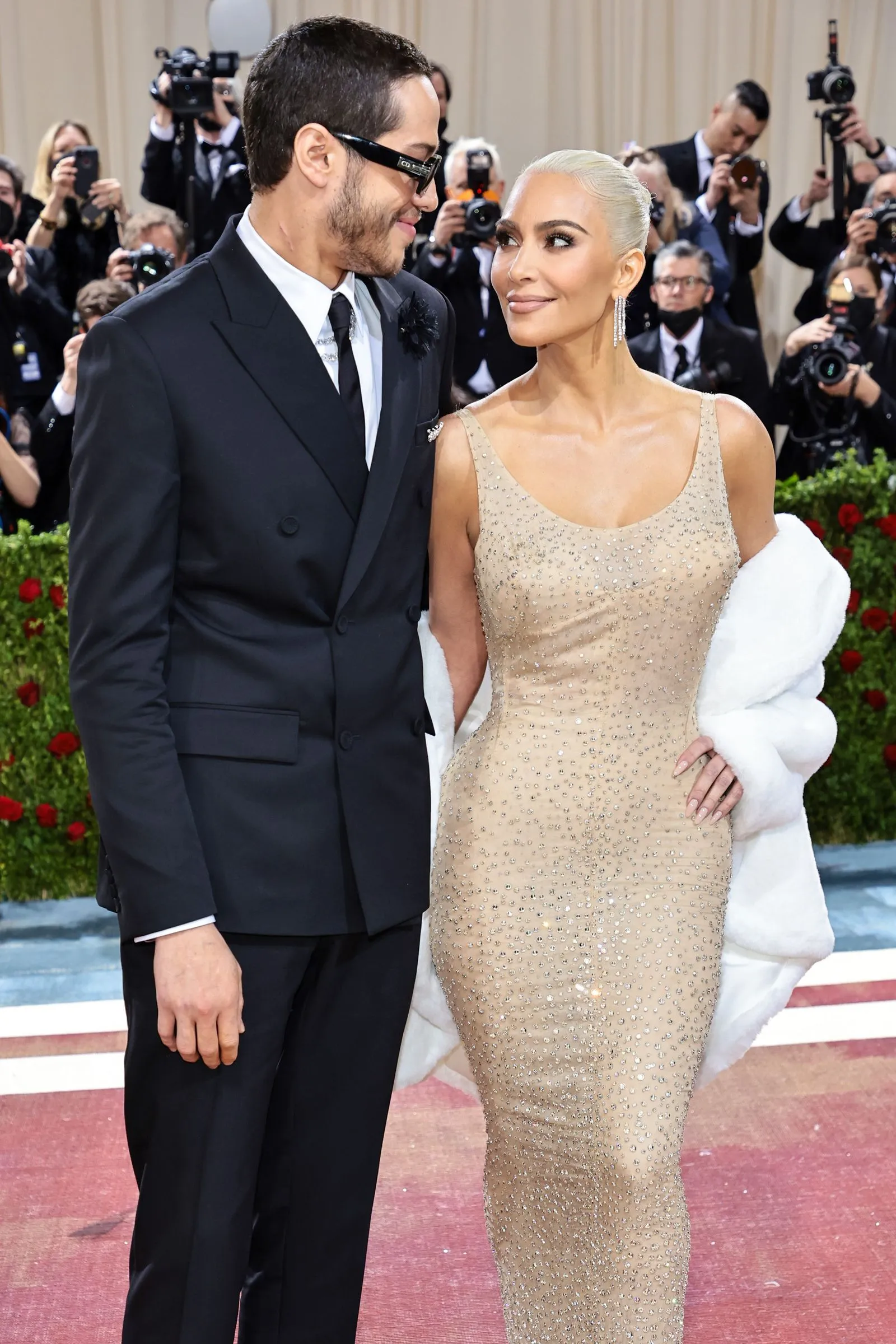 Man in a black suit and sunglasses stands beside a woman in a beige beaded gown on a red carpet, photographers in the background.