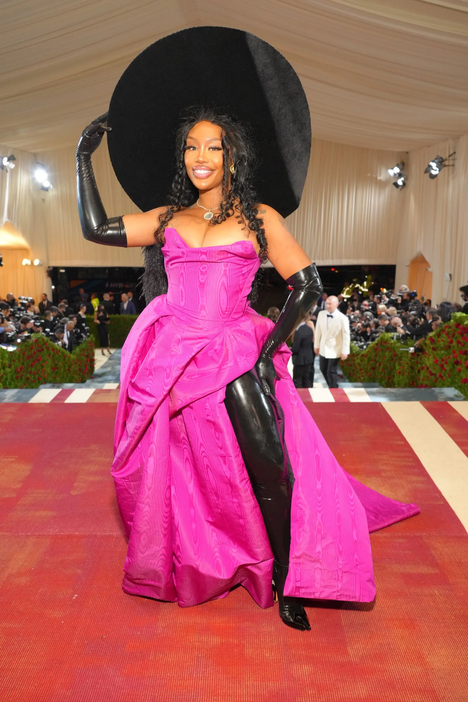 Woman posing on a red carpet in a bright pink ball gown with an oversized black hat and long black gloves, smiling for photographers.