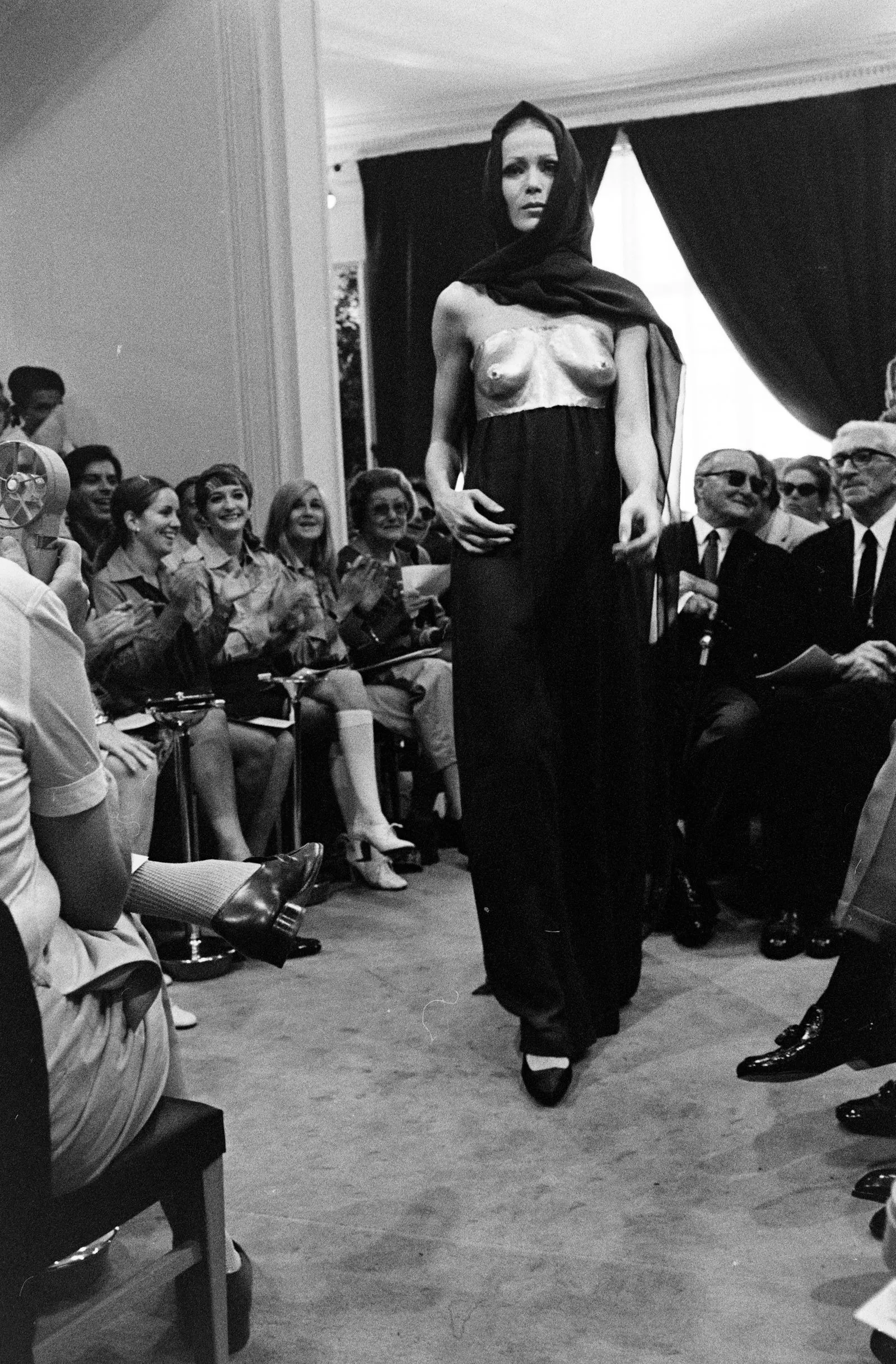 Model walks a runway in a hooded shawl over a metallic strapless top; seated audience applauds.