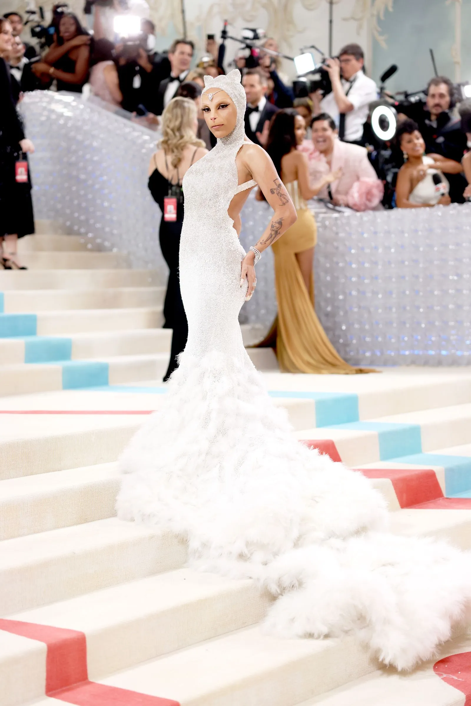 Model in a sparkling white gown with a feathered train posing on a glossy stair carpet at a red-carpet event, paparazzi in the background