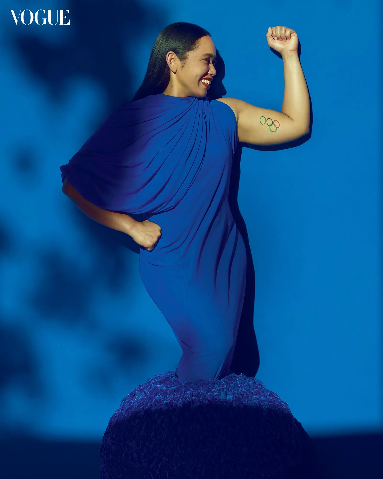 Woman in a blue dress flexes her arm against a blue backdrop, with the Vogue logo in the top-left corner and an Olympic rings tattoo on her upper arm.