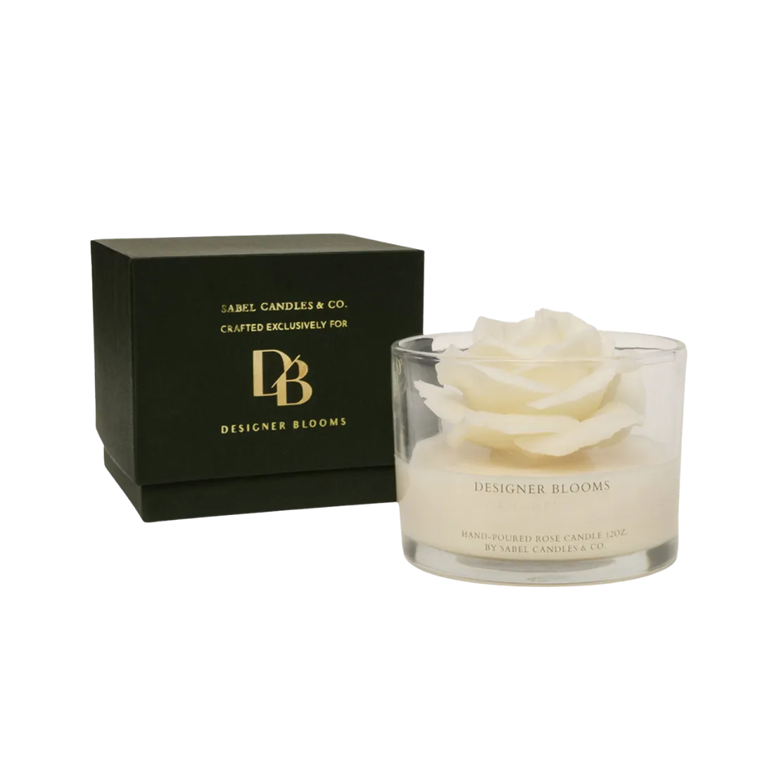 Glass candle jar with a white rose wax centerpiece, labeled 'Designer Blooms', beside a black box with gold branding on cottony packaging beh инд