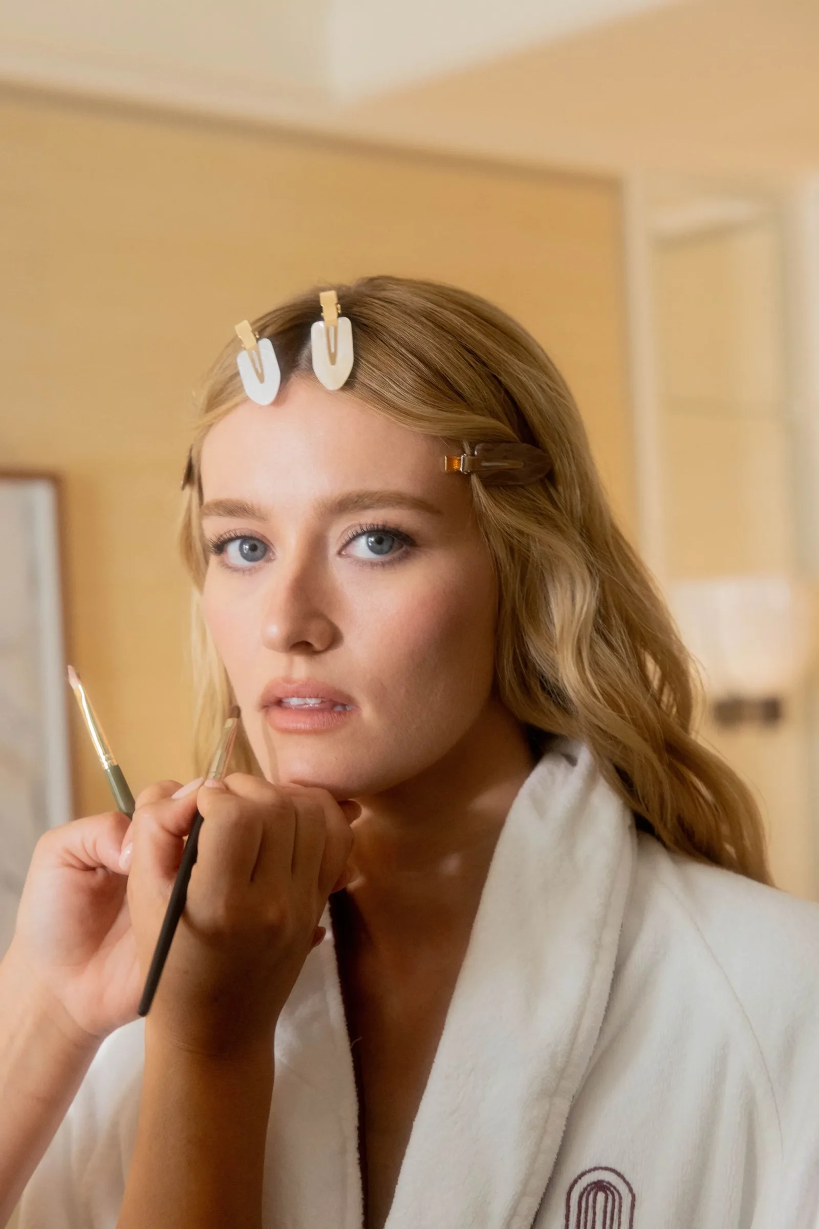 Close-up of a young woman in a white bathrobe applying makeup with a brush, hair clipped back with white clips.