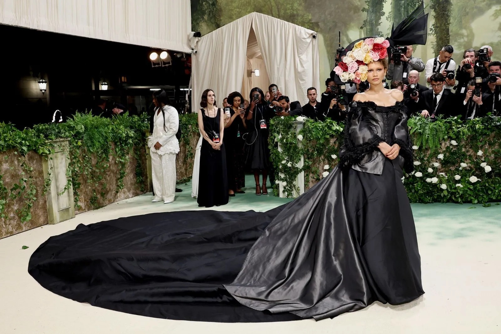 Model in an off-the-shoulder black satin gown with an enormous train and a vibrant flower headdress posing for photographers on a gala carpet.