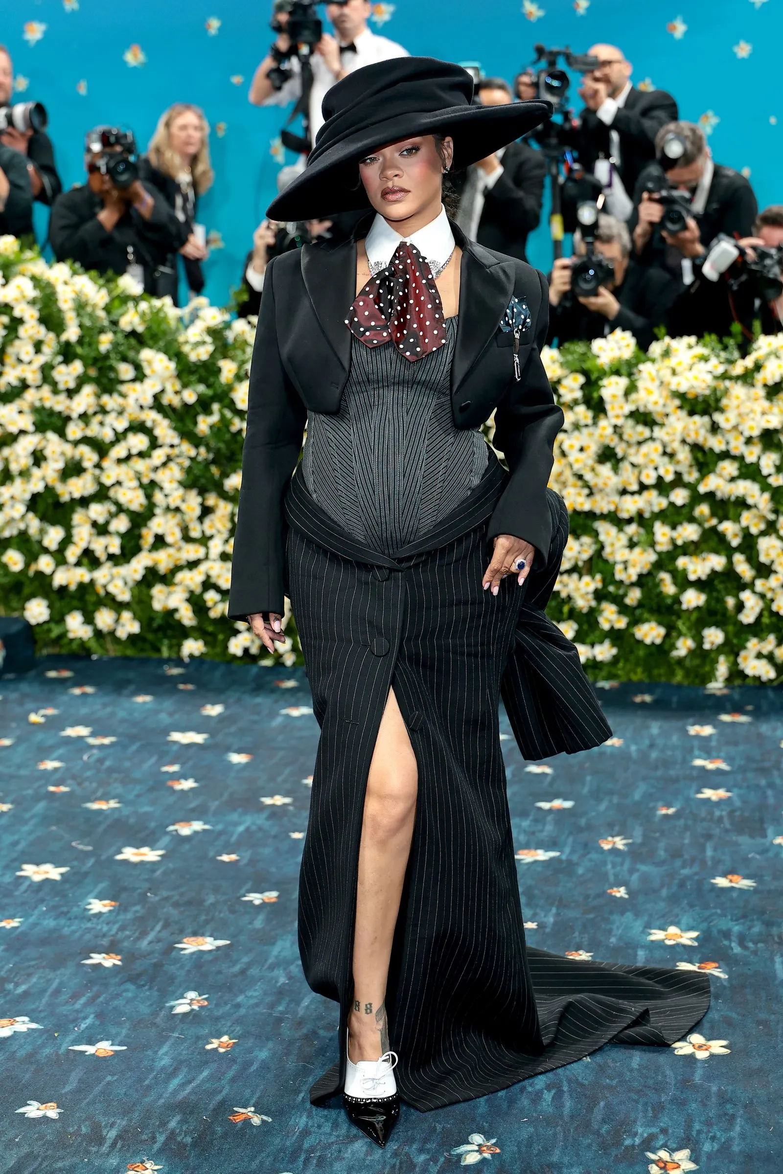 Woman on a blue floral carpet wearing a black pinstripe gown and large black hat, photographers in background capturing the moment.