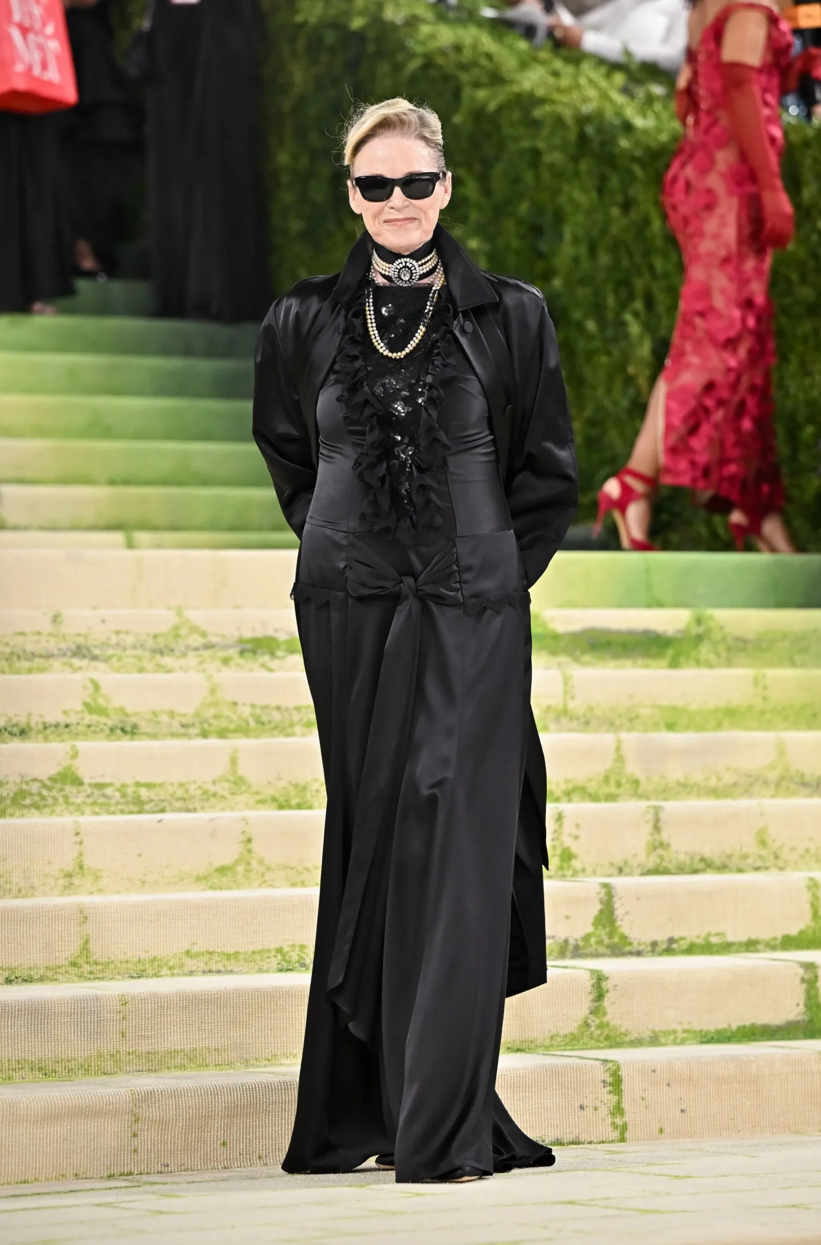 Person in a black satin suit with ruffled blouse and pearl necklace posing on light-colored outdoor steps, wearing dark sunglasses.