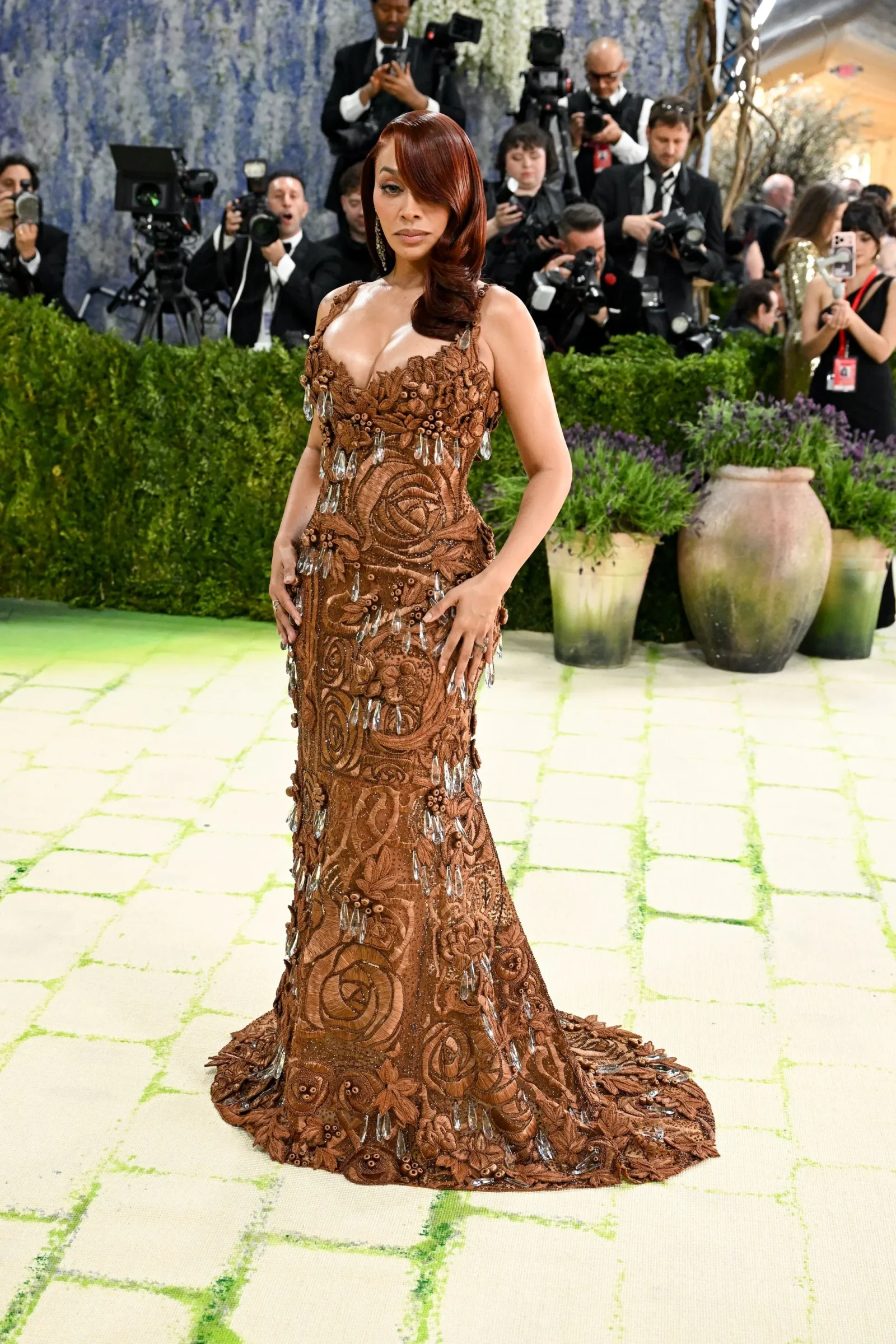 Woman in an ornate brown lace evening gown posing on a green-tiled carpet with paparazzi behind her