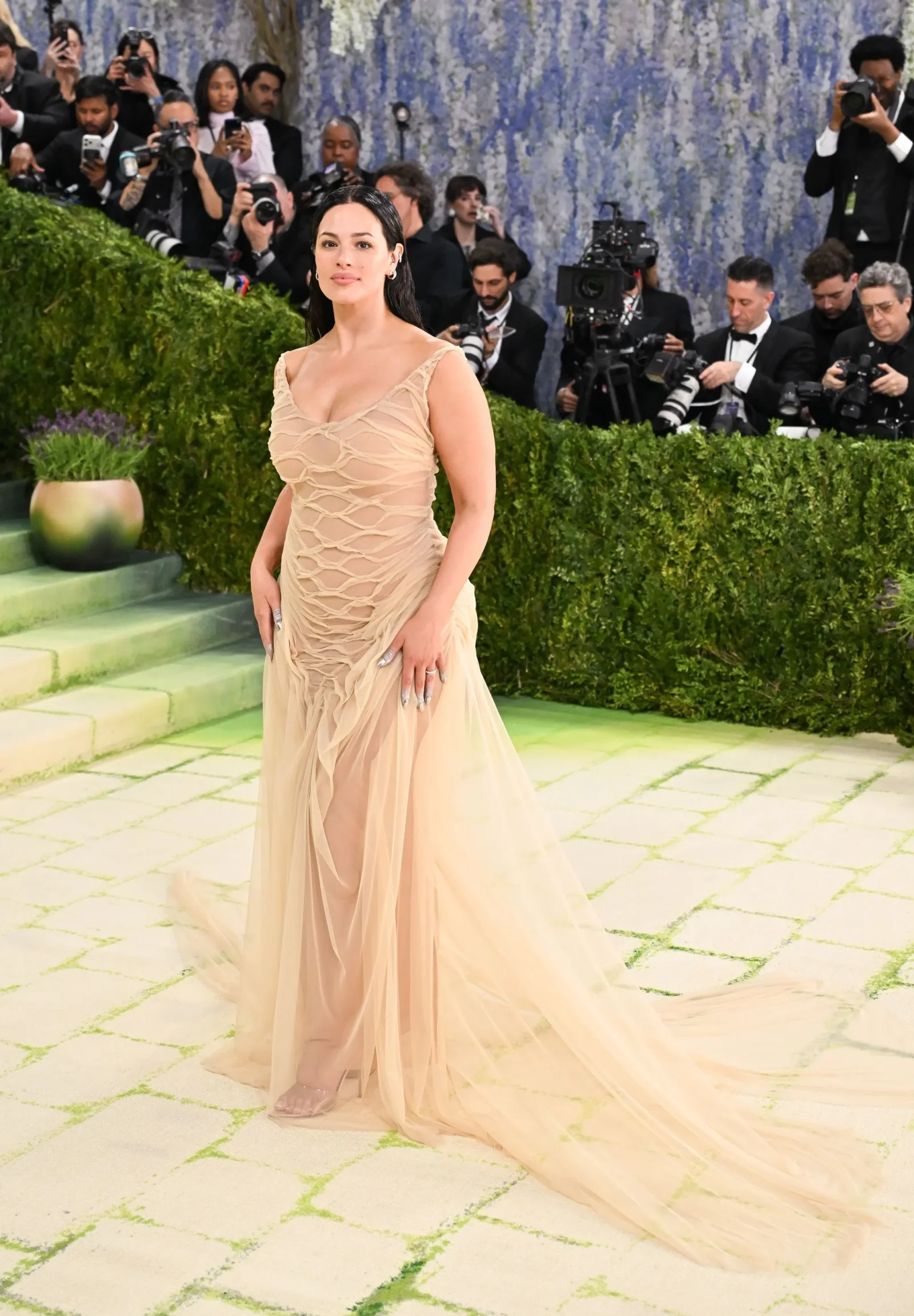 Woman in a beige, sheer, layered evening gown poses on a pale-green carpet as photographers crowd the backdrop behind a hedge.