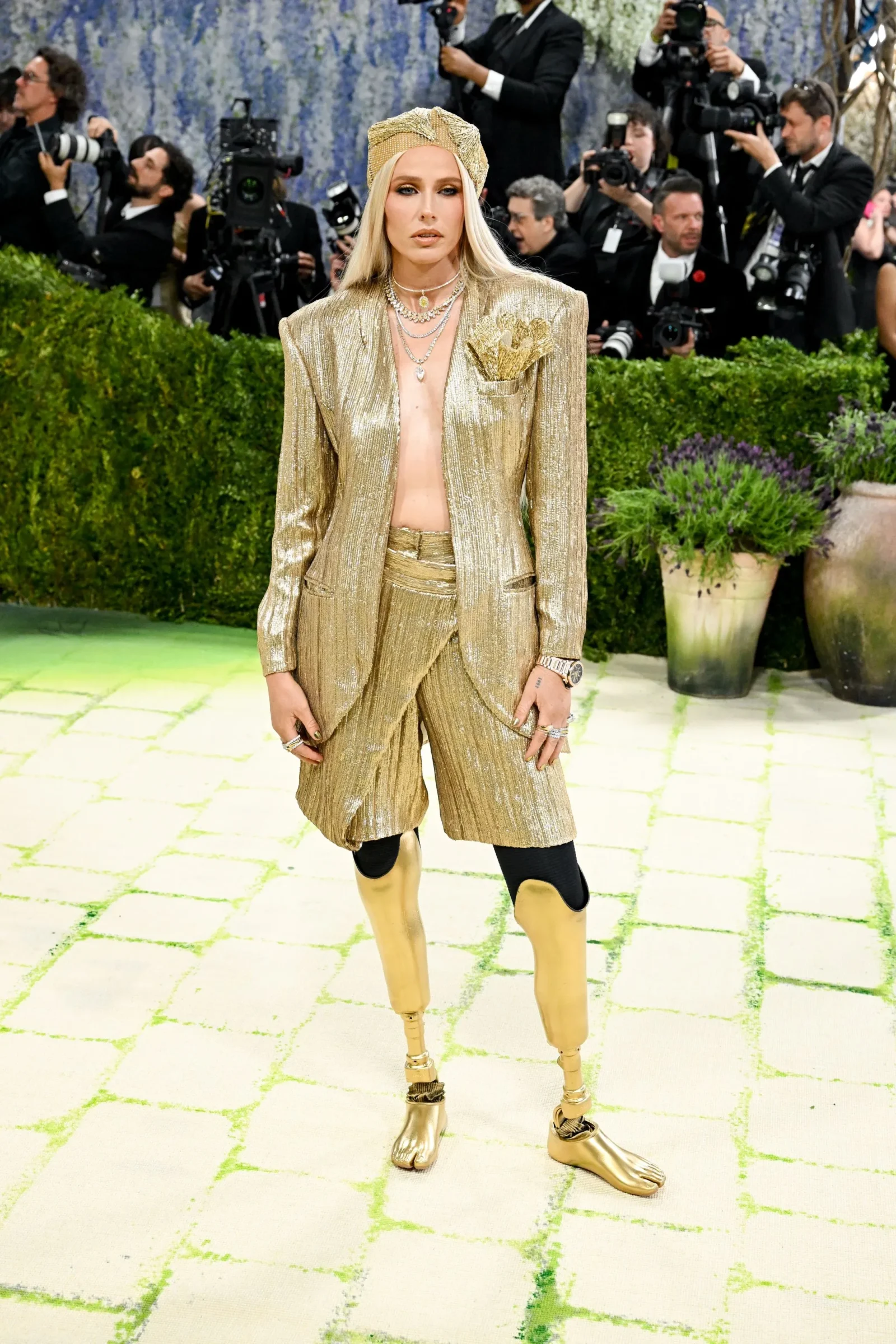 Model wearing a gold metallic suit with matching shorts, head wrap, layered necklaces, and knee-high gold boots at a press event with photographers in the background.