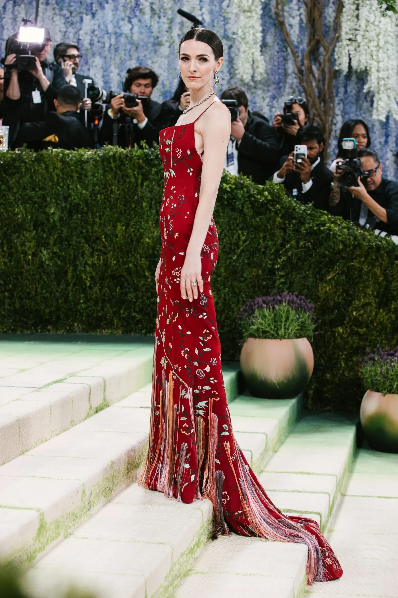 Woman in a red floral evening gown with long fringe stands on white steps at a red-carpet event, photographers in the background.