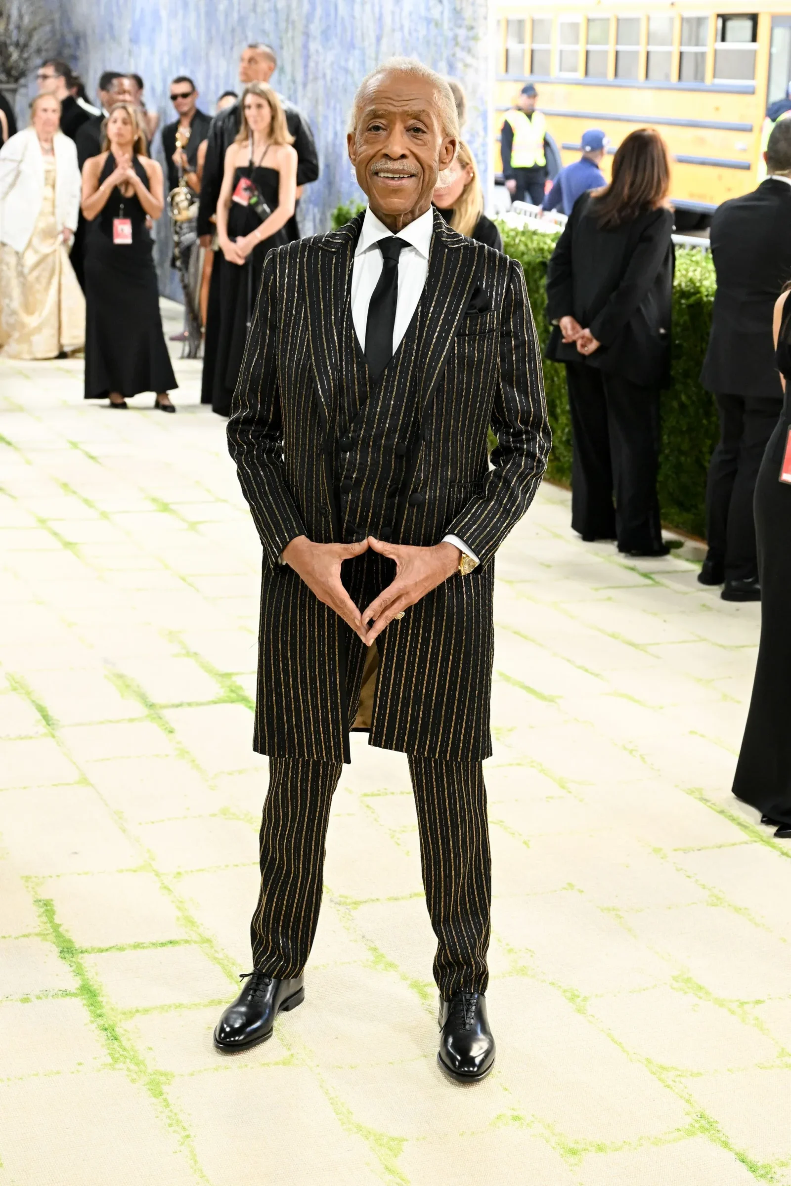 Elderly man in a black pinstripe suit with a white shirt and black tie poses for a photo at a formal event, hands together in front, others in black attire and a yellow bus in the background.