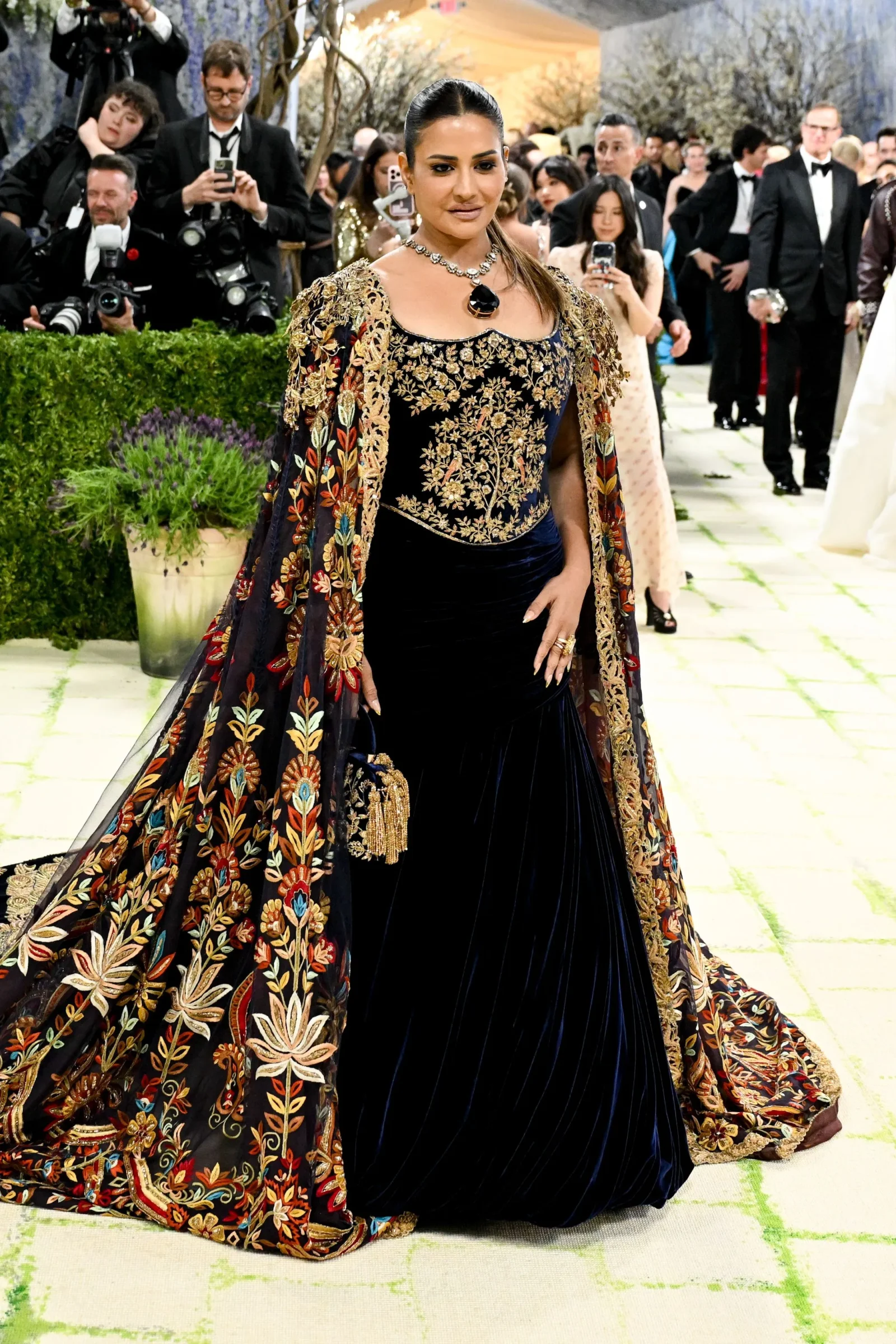 Elegant woman in a dark blue velvet gown with gold-embroidered cape, posing at a formal event as photographers surround her in the background.
