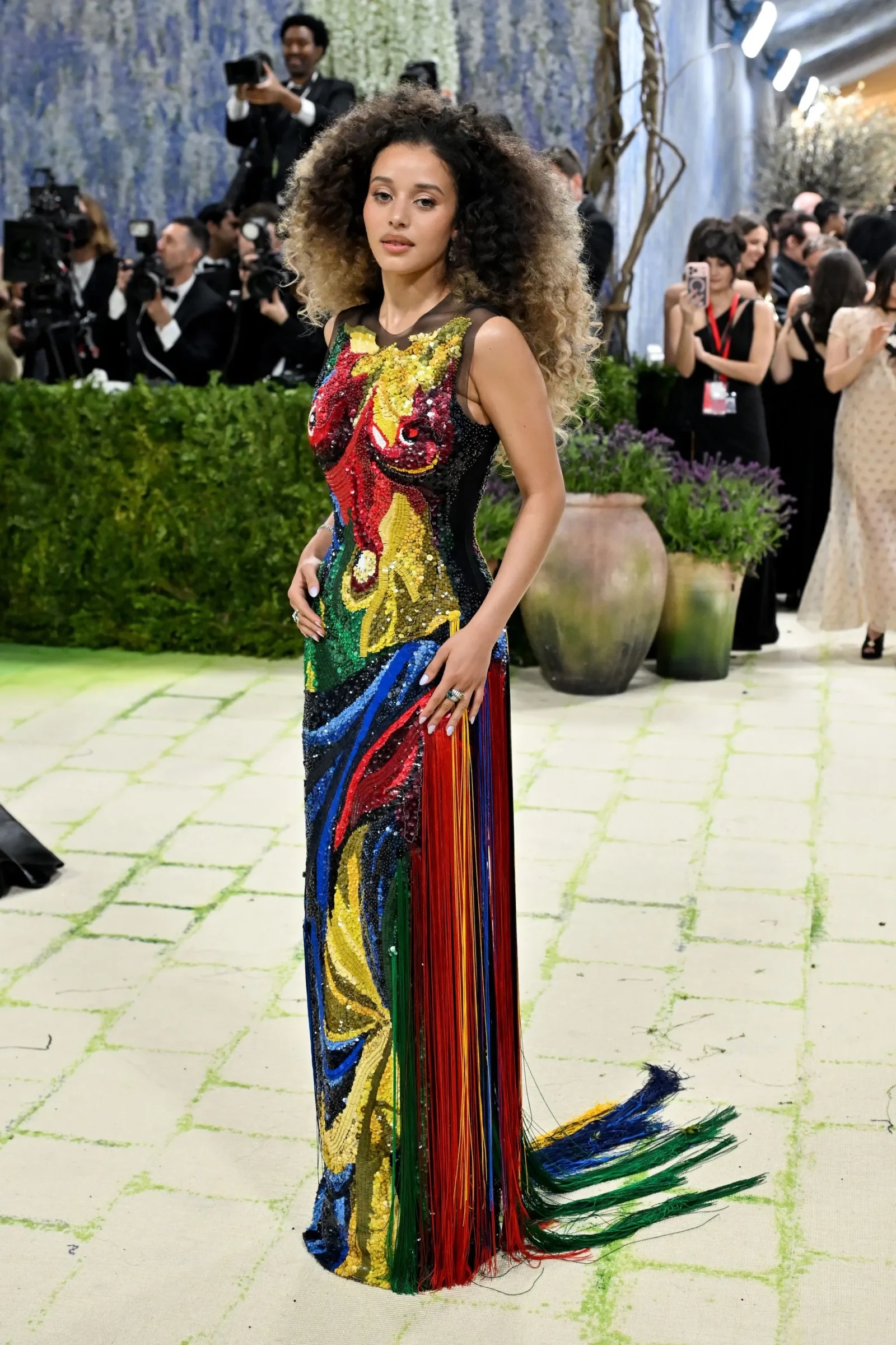 Woman wearing a bright multicolor sequined gown with fringe, posing on a carpet; paparazzi in the background.