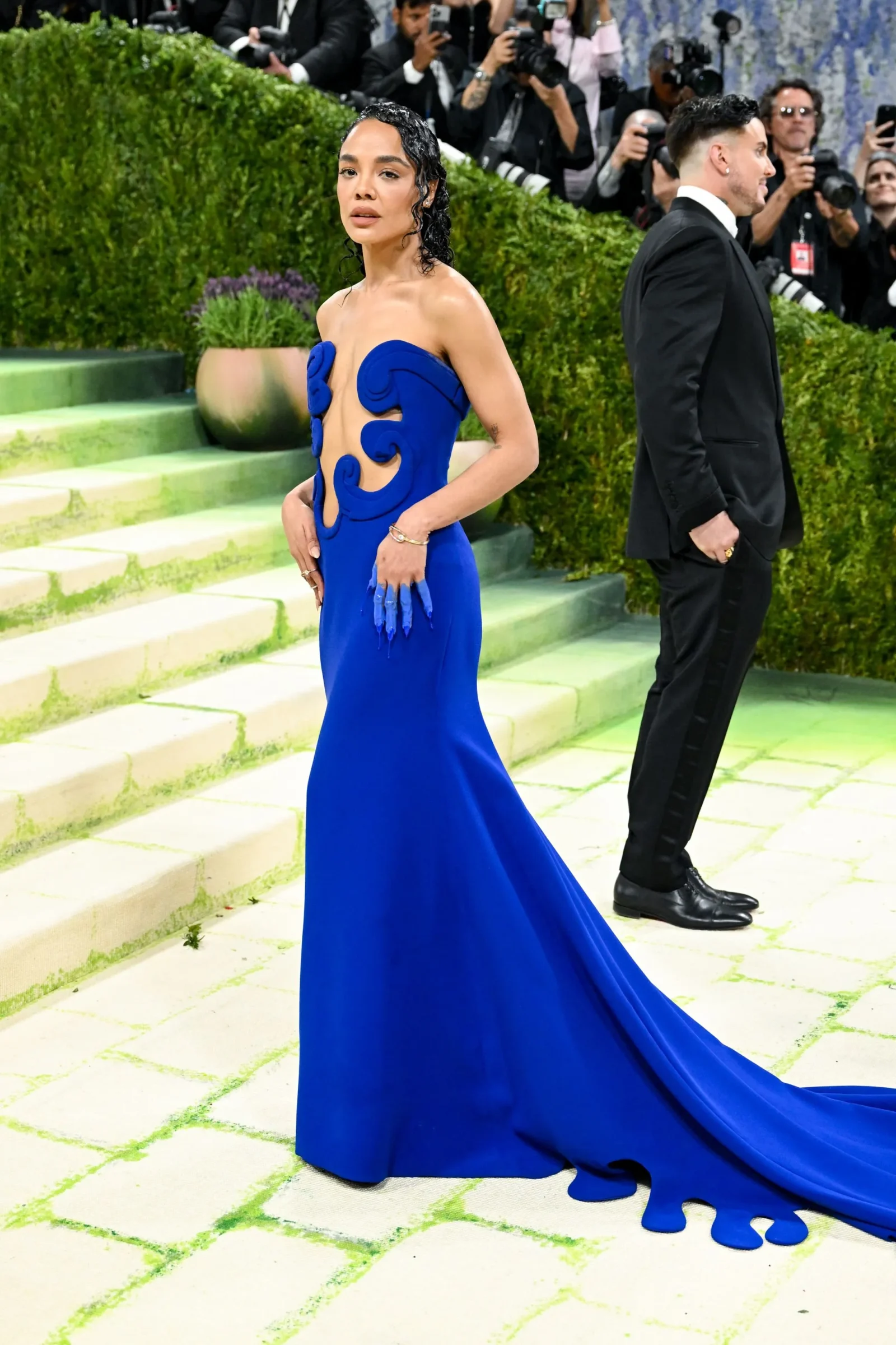 Woman in a royal blue gown with sculpted cutouts posing on lime-green carpeted stairs as photographers document the moment.