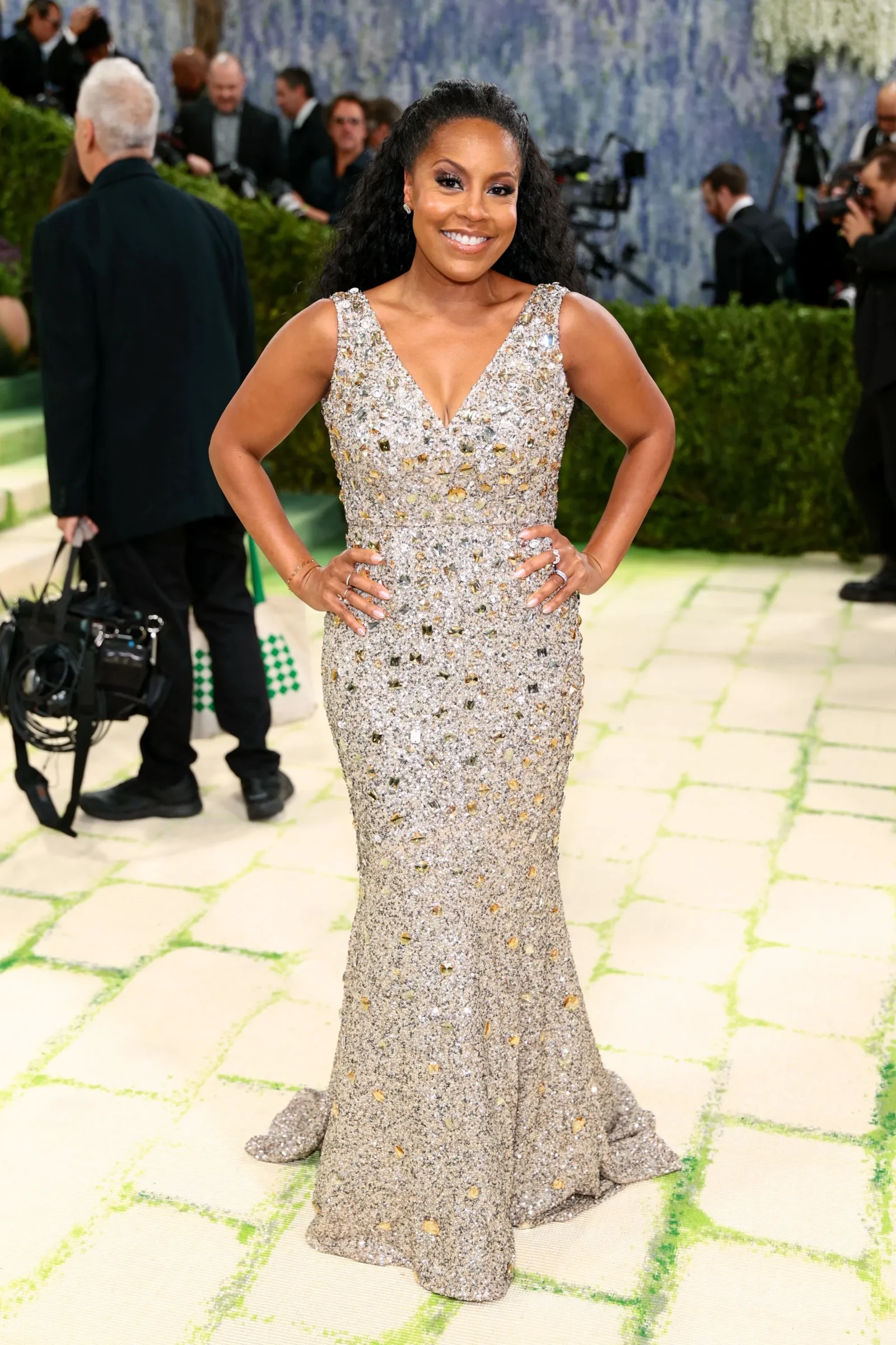 Woman posing on a red carpet in a silver embellished gown, with photographers in the background and a green backdrop.