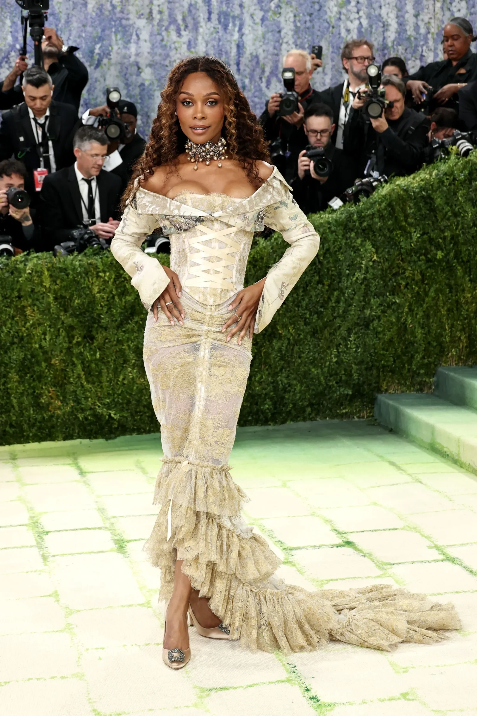 Woman in an off-shoulder gold gown with corseted waist posing on a runway as photographers flash in the background.