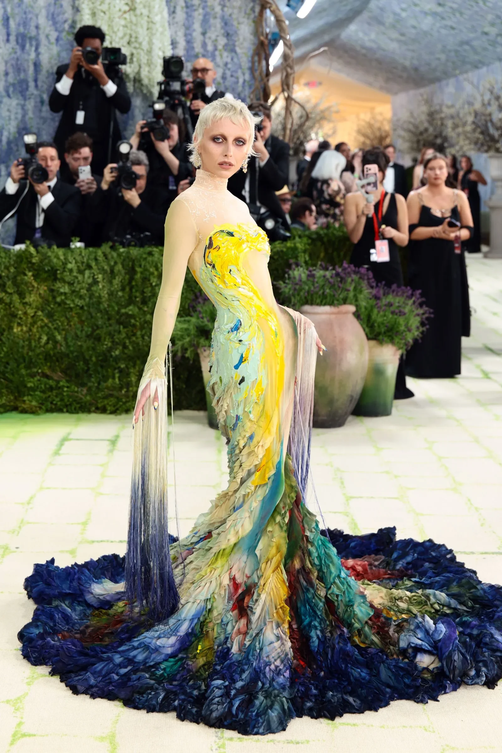 Model on a runway wearing a colorful, fringe-trimmed gown with a long train; photographers line the background taking photos.