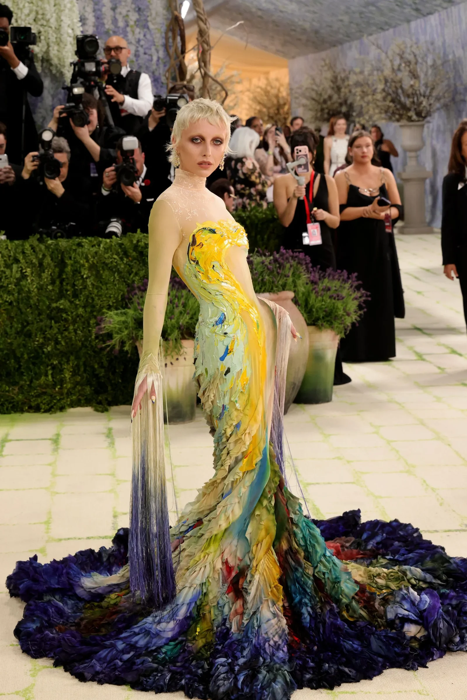Model wearing a colorful, flowing gown with fringe trains on a patterned runway, photographers in the background capture the moment.
