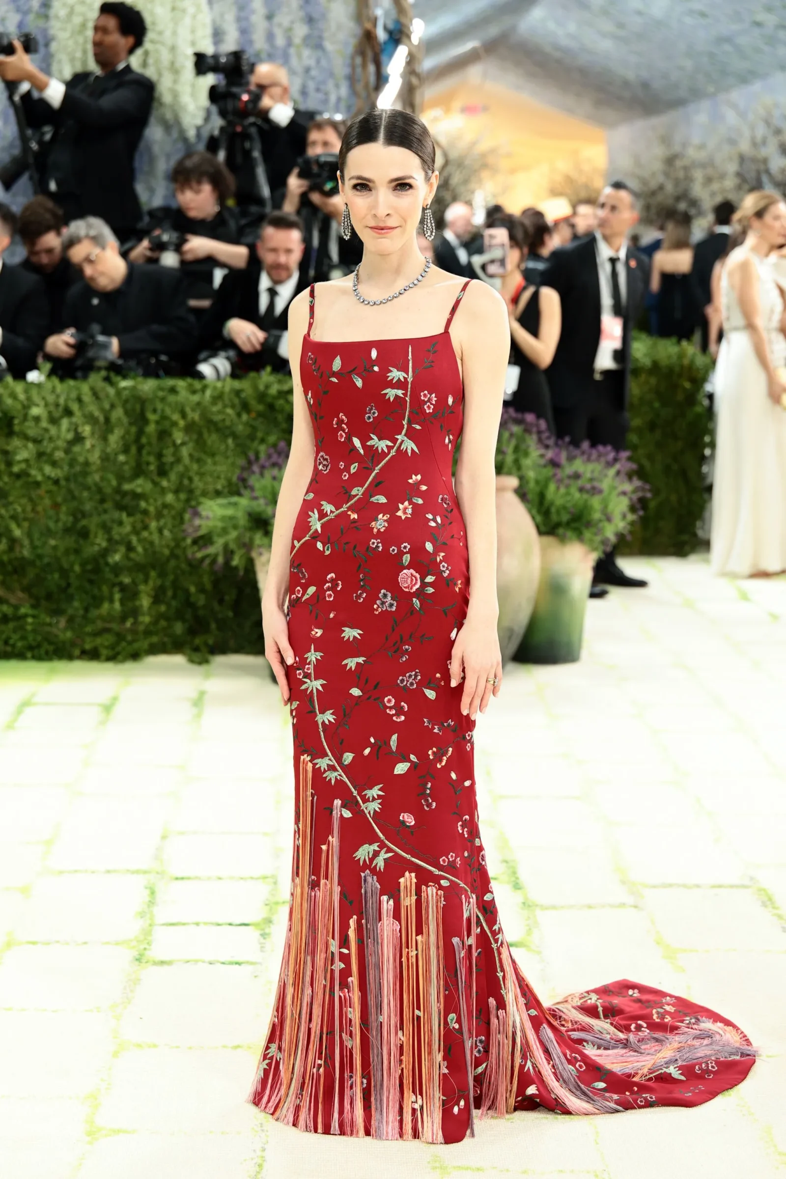 Portrait of a woman in a red floral gown posing on a red carpet with photographers in the background.