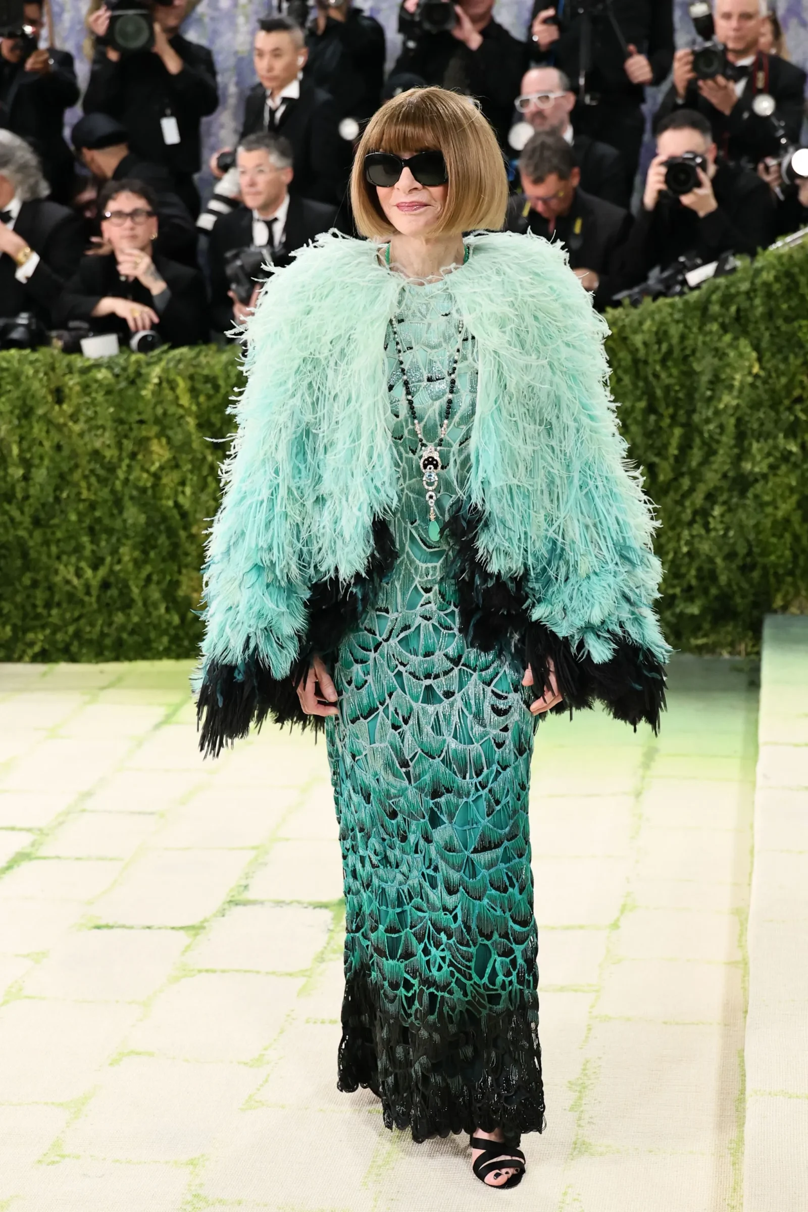 Woman wearing a turquoise feathered cape over a patterned green gown walks the red carpet as photographers shoot nearby, black sunglasses on and jewelry visible.