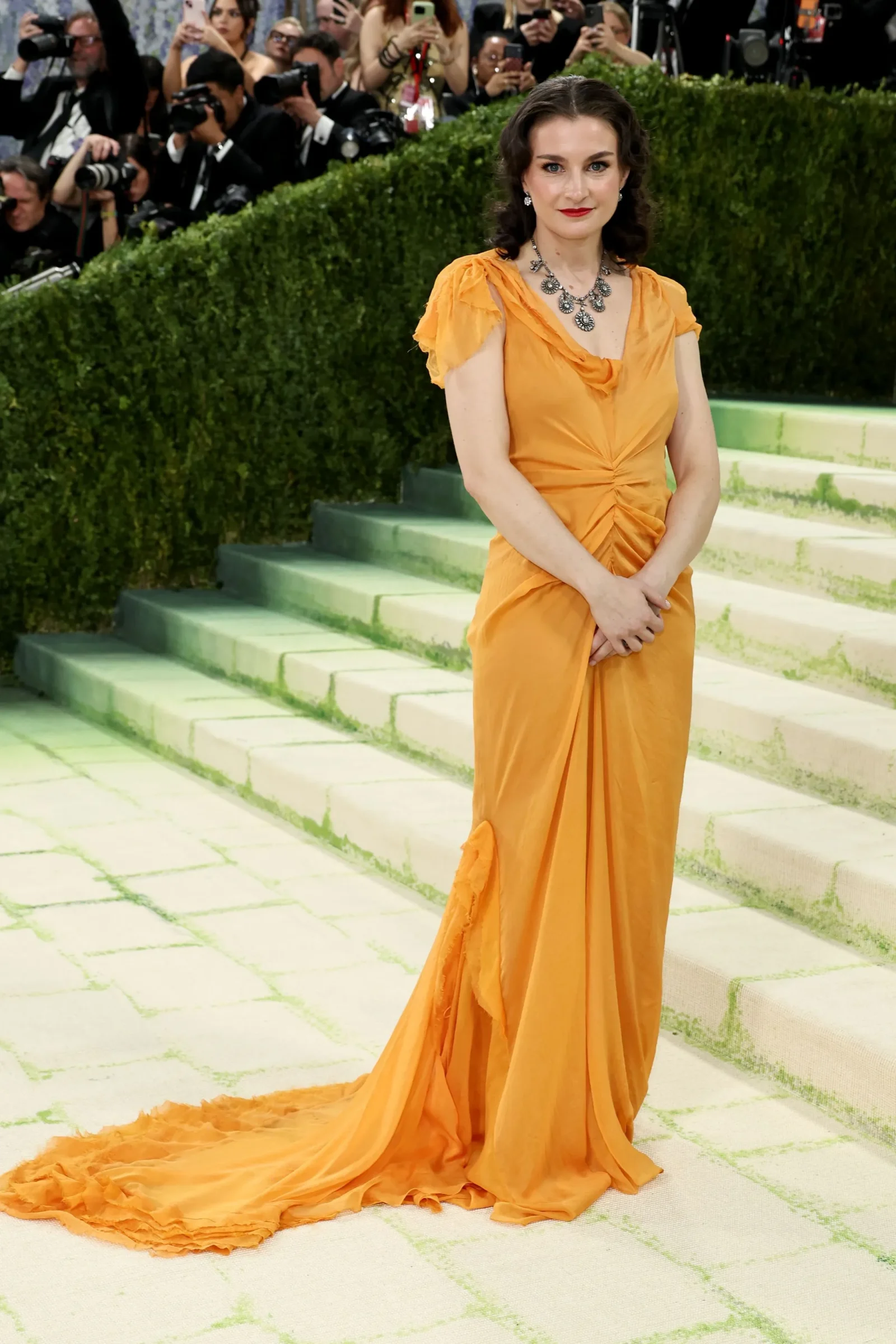 Elegant woman in a bright orange gown with a flowing train posing on steps at a red-carpet event, photographers in the background.