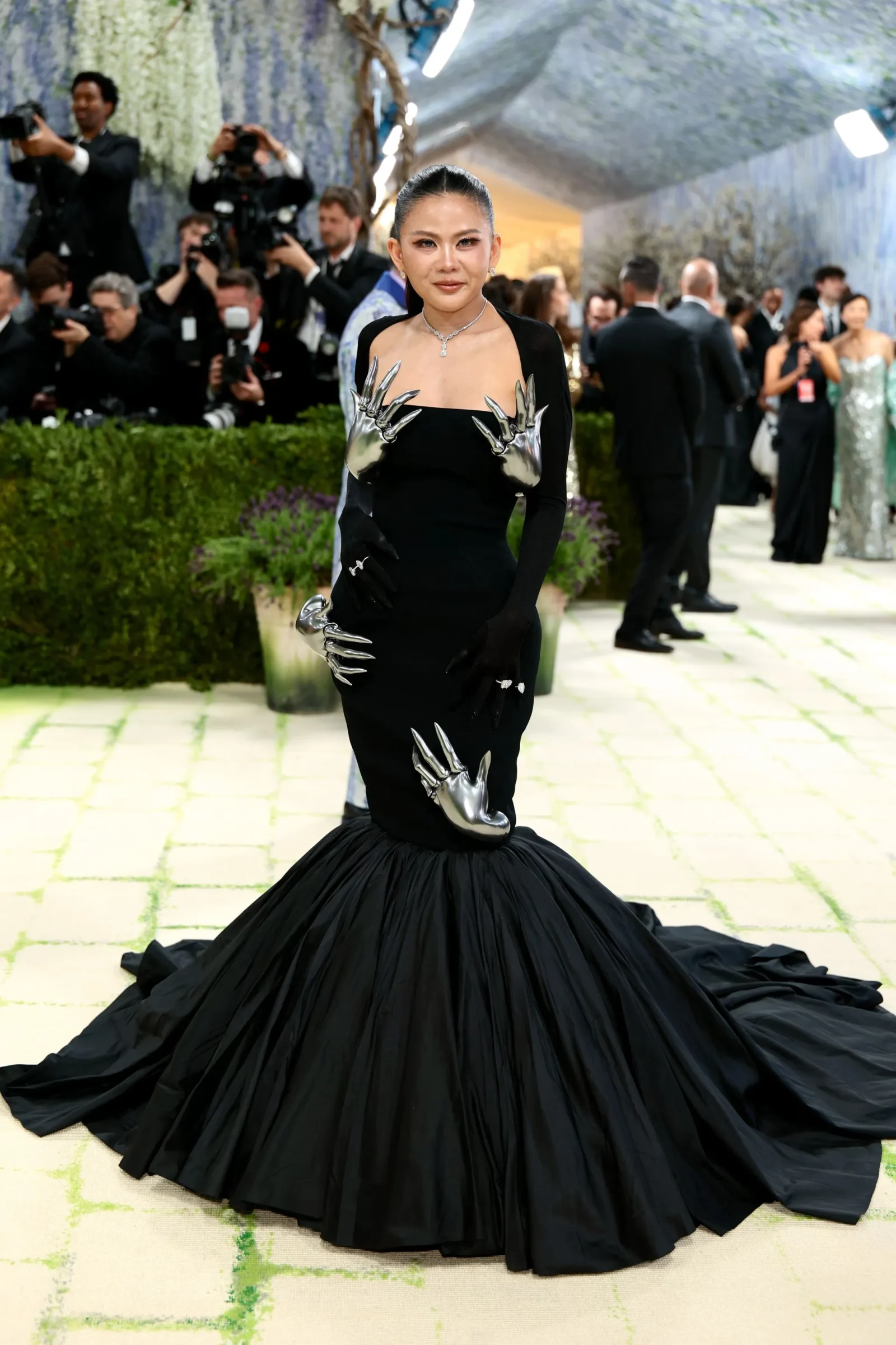 Woman on a red carpet wearing a black gown with silver claw-like sleeves and a long train, posing for photographers.