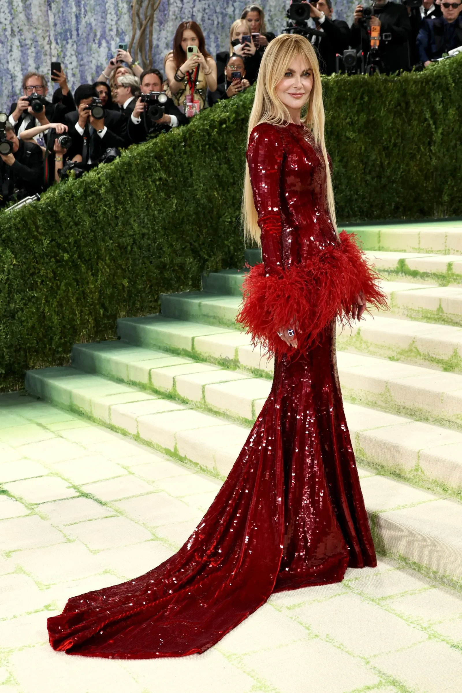 Woman in a floor-length red sequined gown with feathered cuffs posing on light-colored stairs at a red carpet event, photographers in the background taking photos.